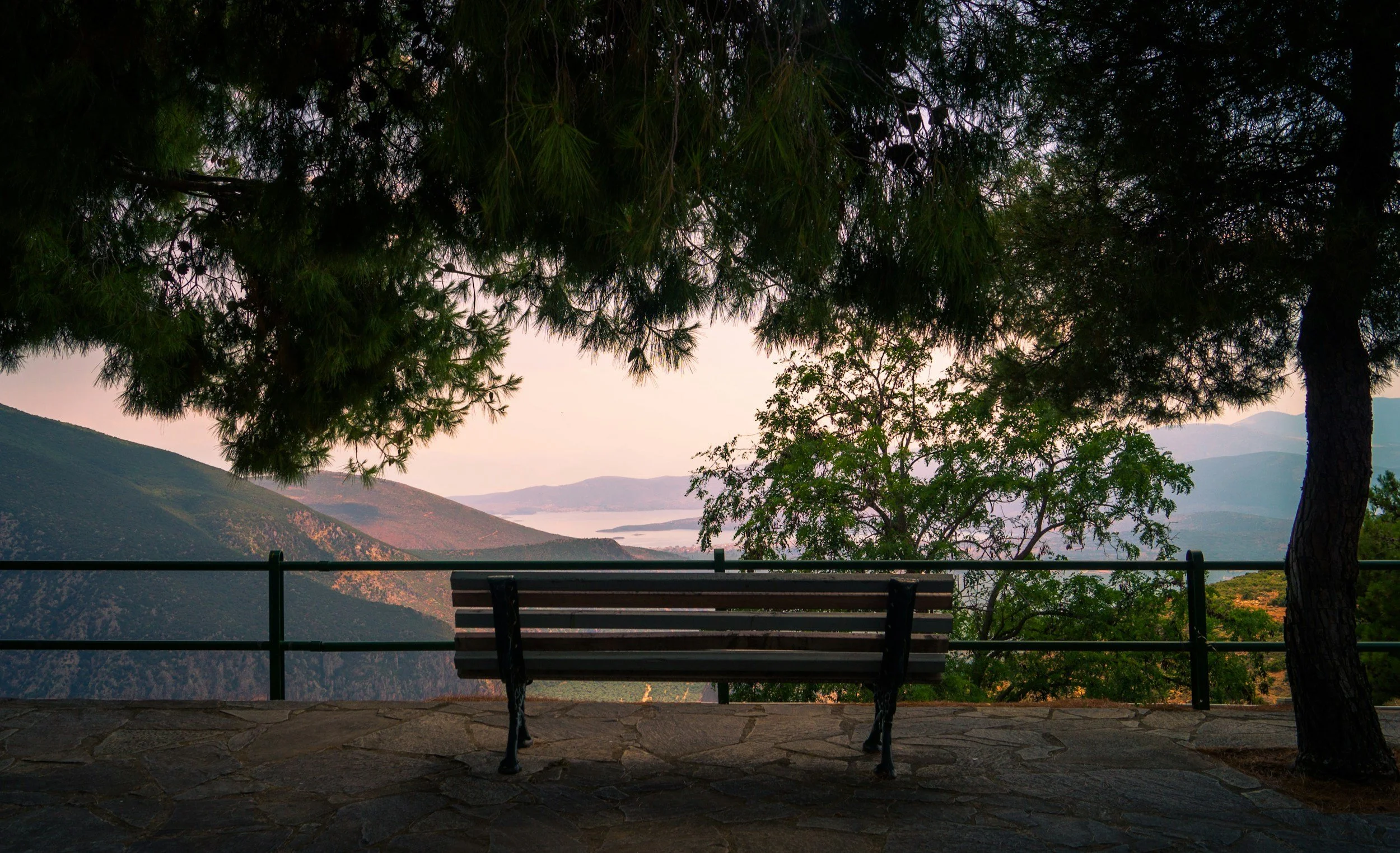 A scenic view of mountains and a body of water seen through a canopy of pine trees with a park bench and a railing in the foreground.