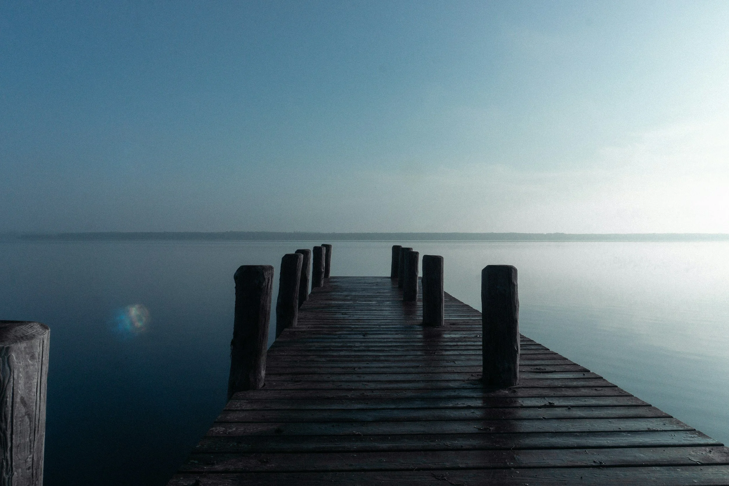 A wooden dock extending into a calm body of water under a clear, pale sky with soft light.