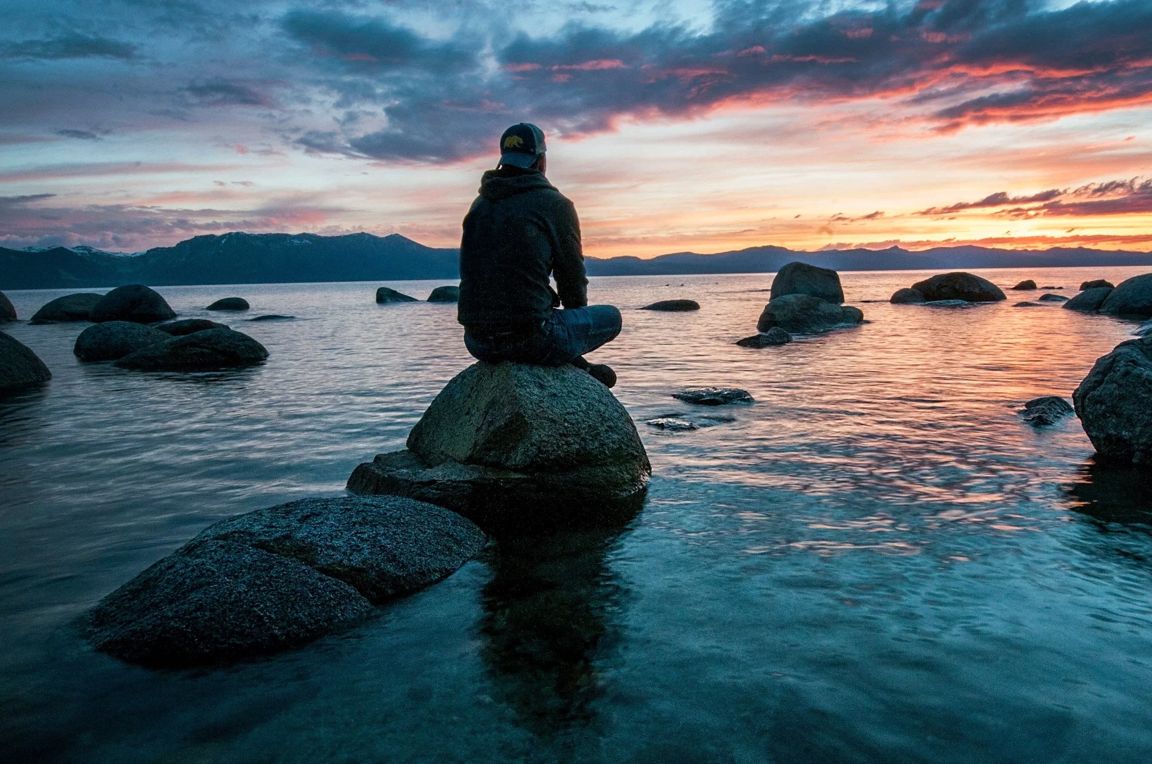 A person sitting cross-legged on a large rock by a lake during sunset, with mountains in the background and a sky filled with colorful, cloudy streaks.