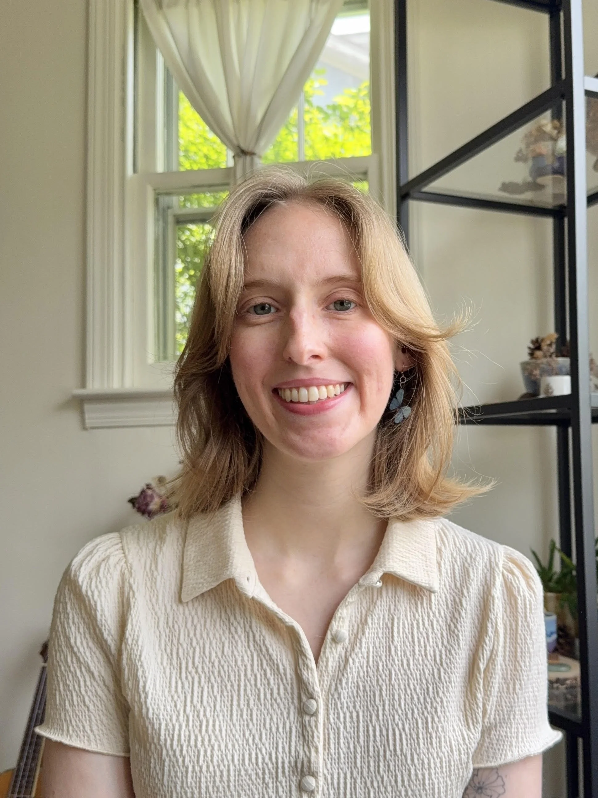 A woman with shoulder-length blonde hair smiling, wearing a cream-colored textured button-up shirt, standing indoors with a window and greenery in the background.