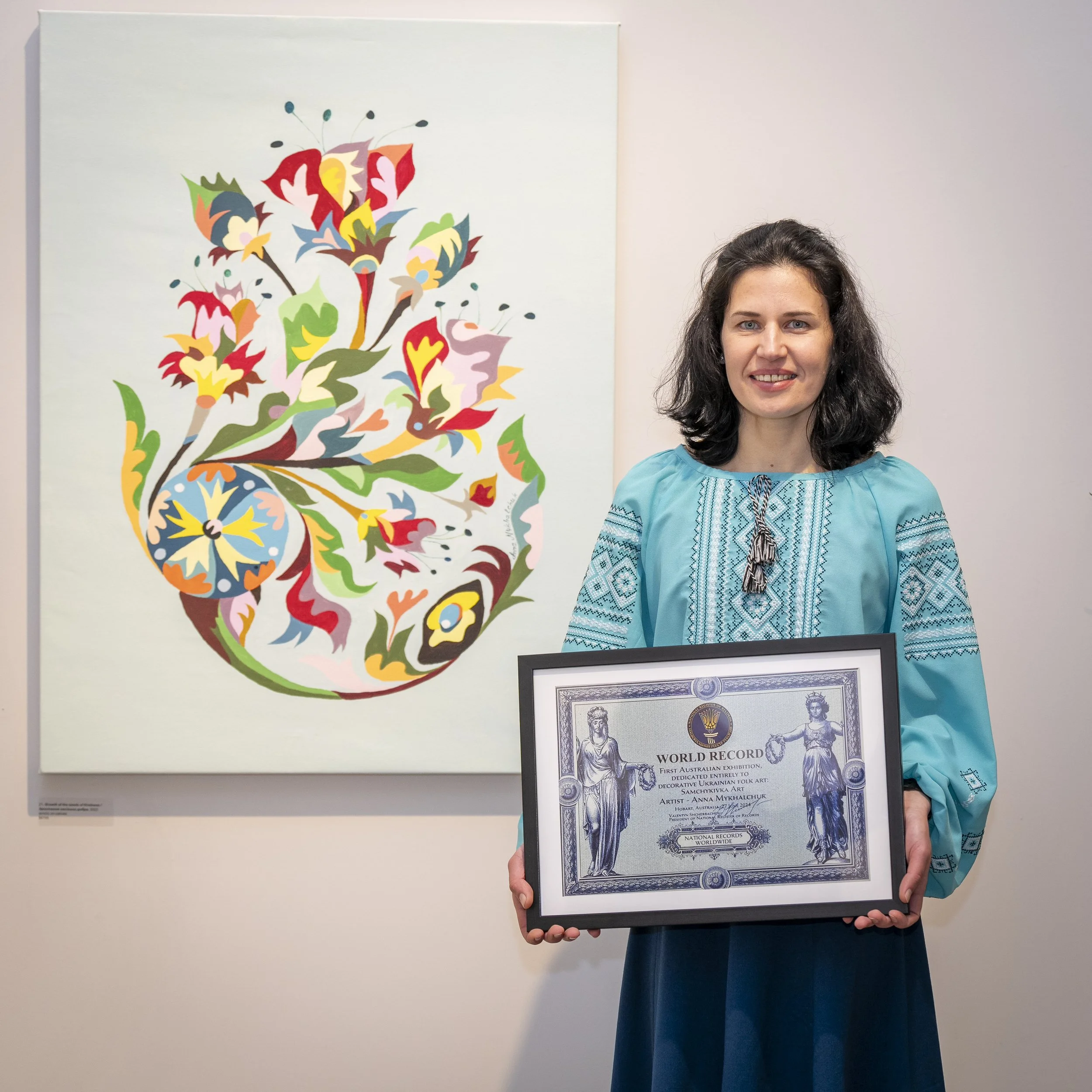 Anna Mykhalchuk in blue traditional dress holding a framed certificate in front of colorful abstract Samchykivka artwork at het  exhibition at Australia.