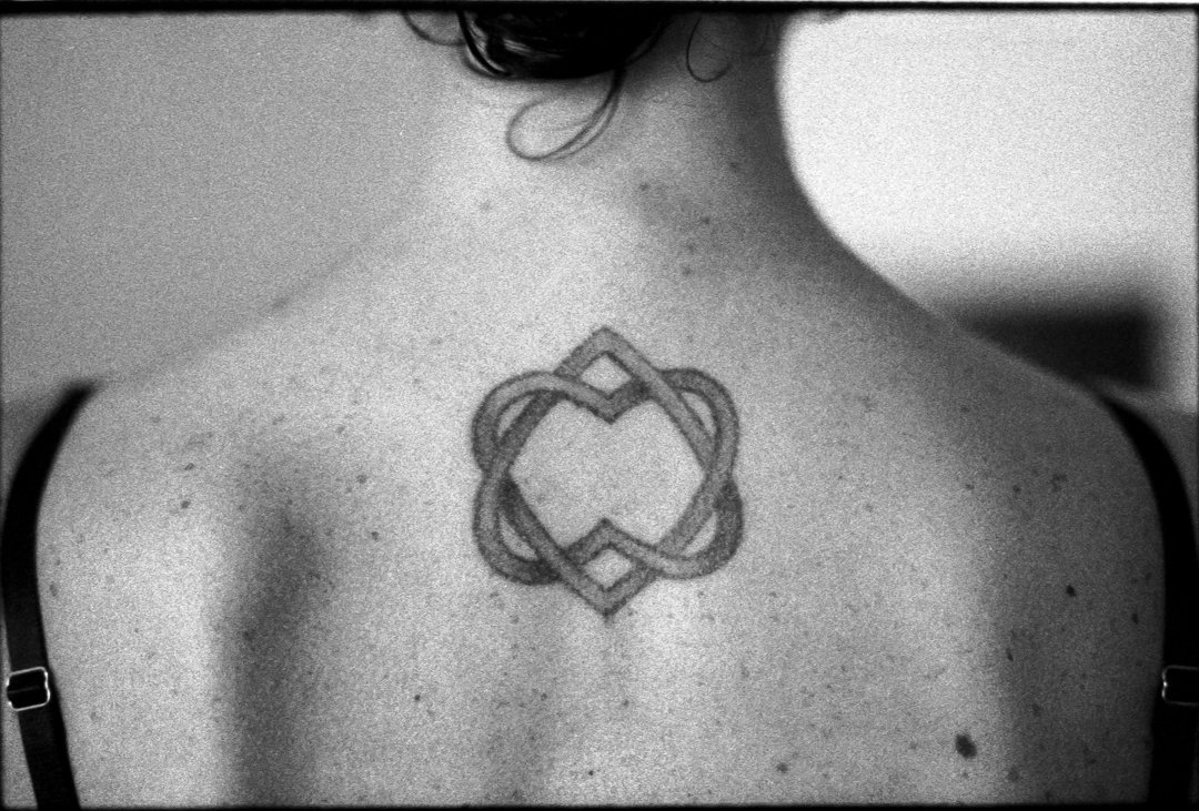 A black and white photo of a person's upper back and shoulder, showing a geometric tattoo with intertwined lines forming a symmetrical design.