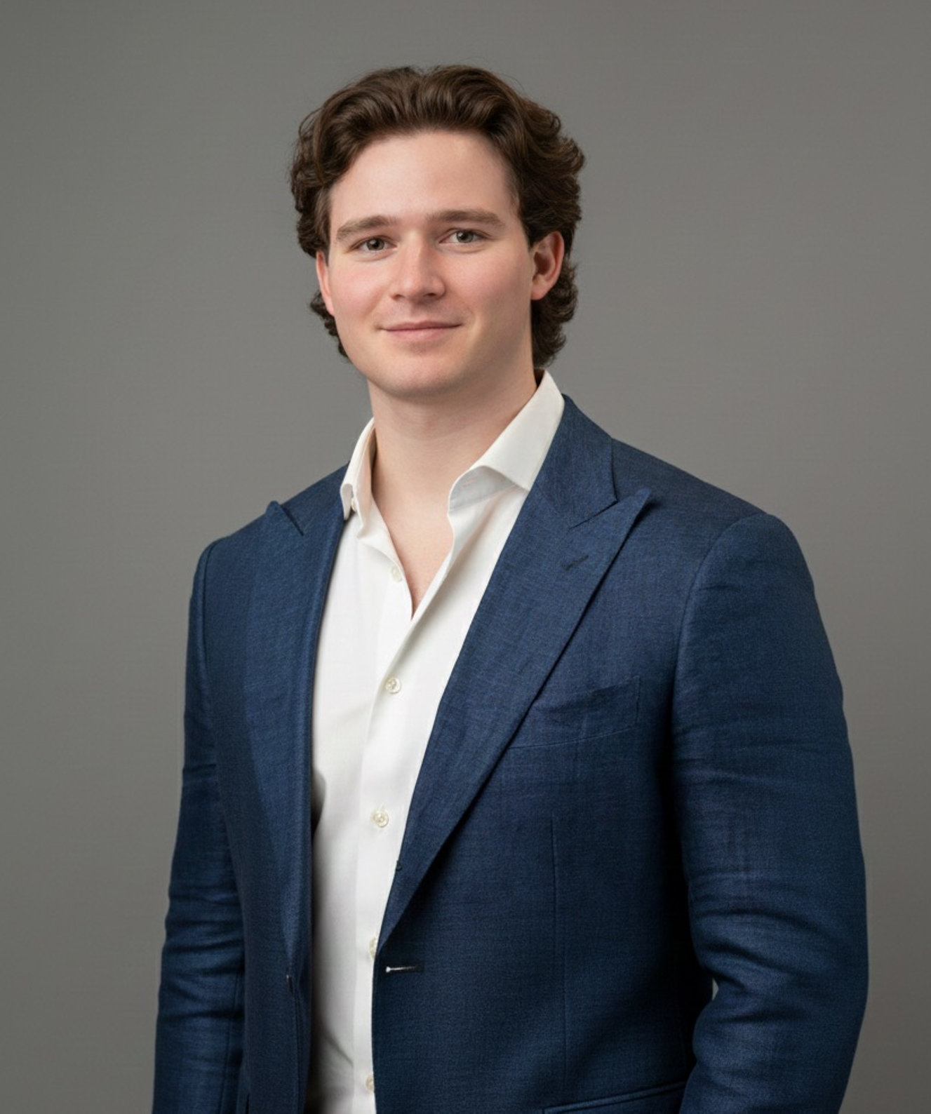 A young man in a dark suit, white shirt, and striped tie standing outdoors in front of a tree with greenery.