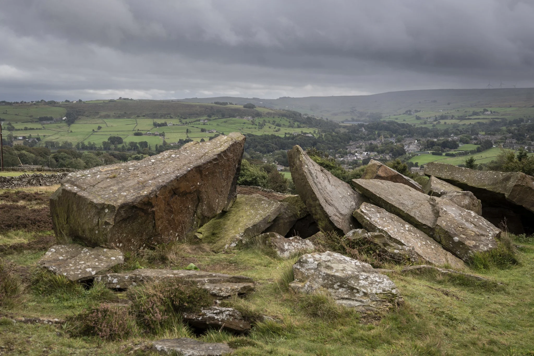 Large rocks in a grassy field with a countryside landscape in the background, featuring rolling hills and a cloudy sky.