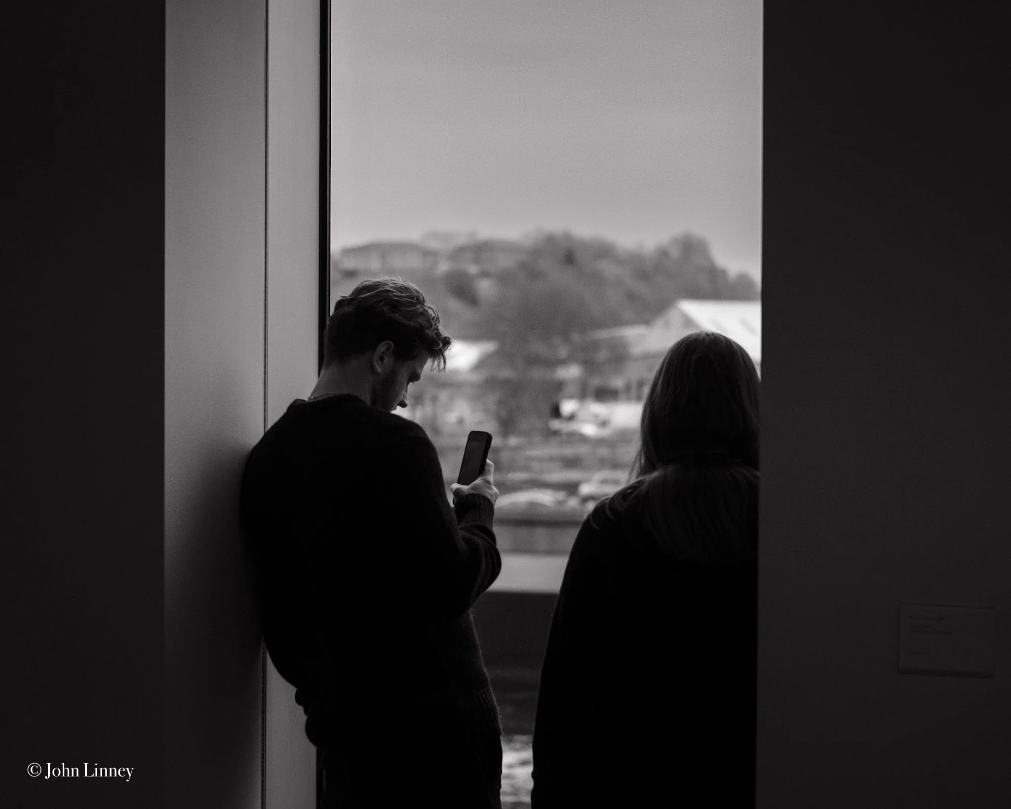 Silhouetted people standing by a window, one looking at a phone, with an outdoor urban landscape in the background.