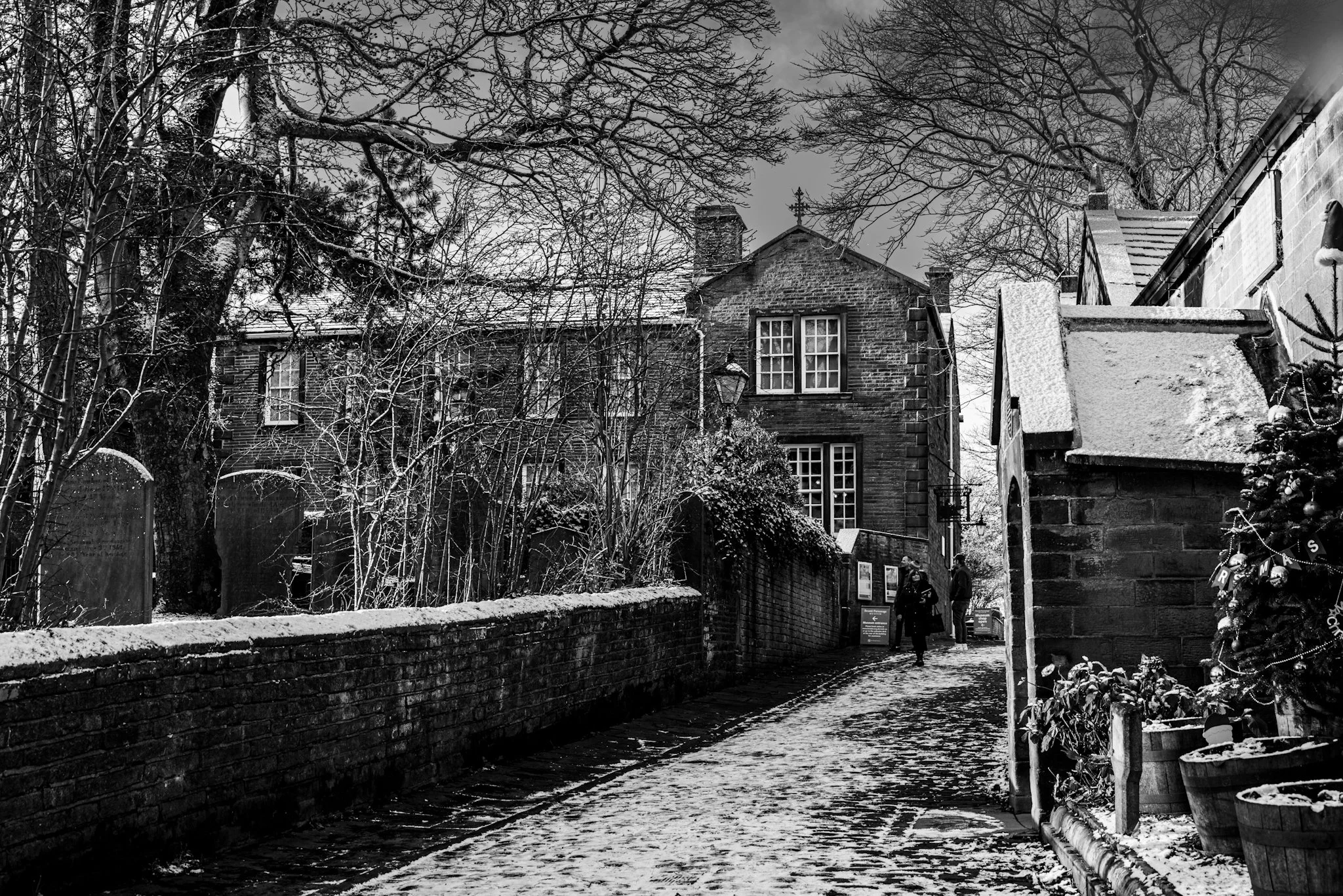 Cobblestone path in a historic village with old brick buildings, snow on the ground and rooftops, bare trees, and a Christmas tree with ornaments.