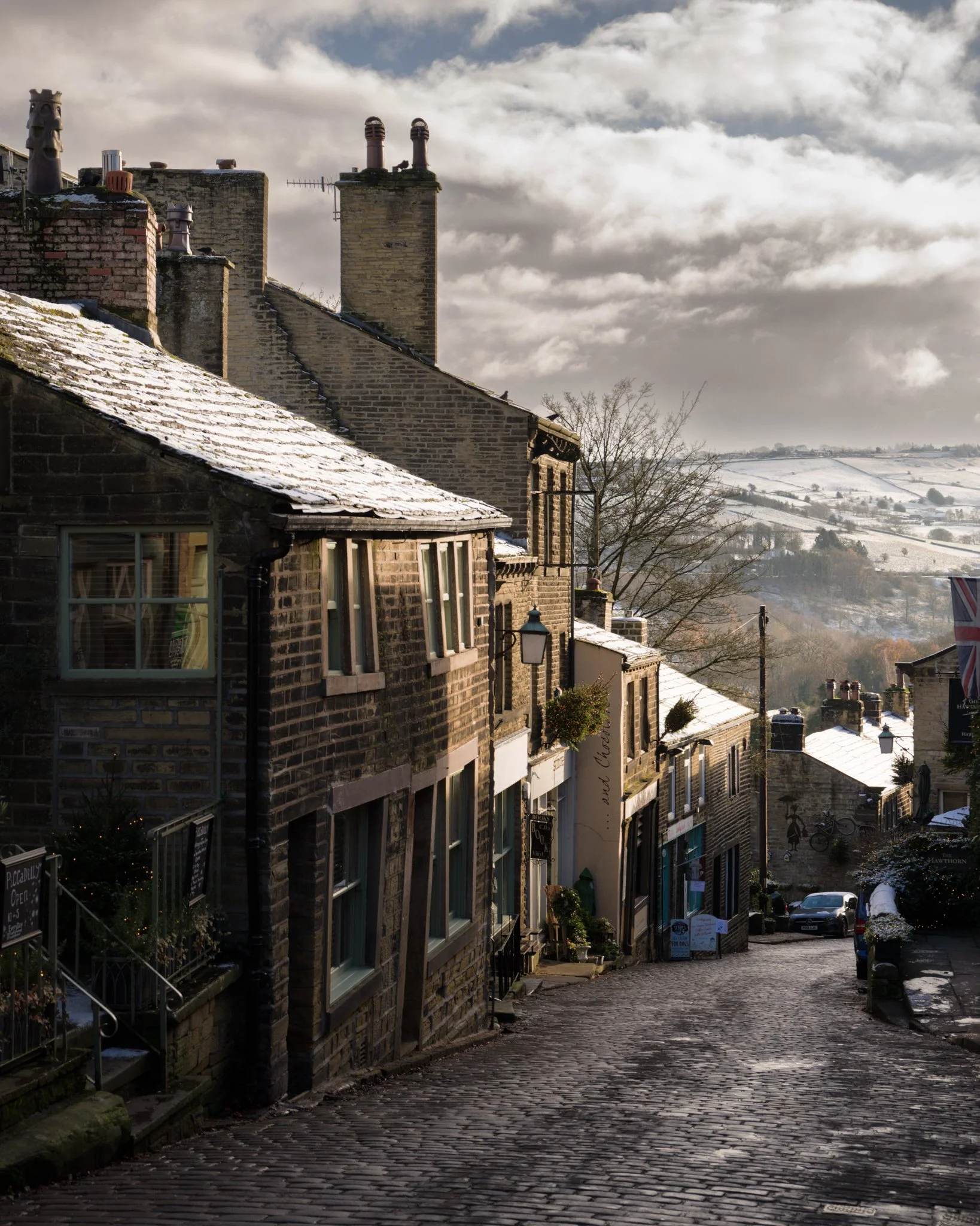 Steep cobblestone street with stone buildings and a snow-dusted rooftop, leading down to a picturesque valley under a cloudy sky.