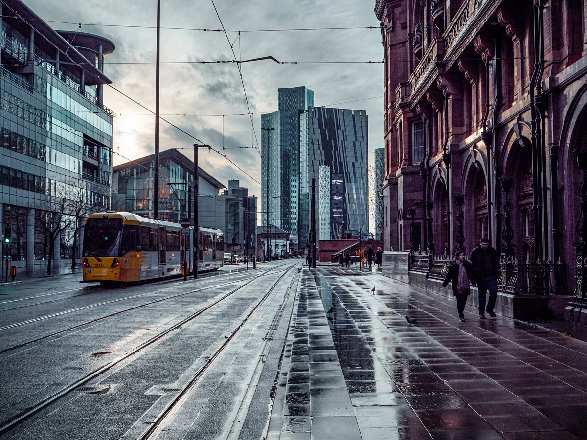 Urban scene with a tram on a city street, surrounded by modern and historic buildings, reflecting on wet pavement after rain.