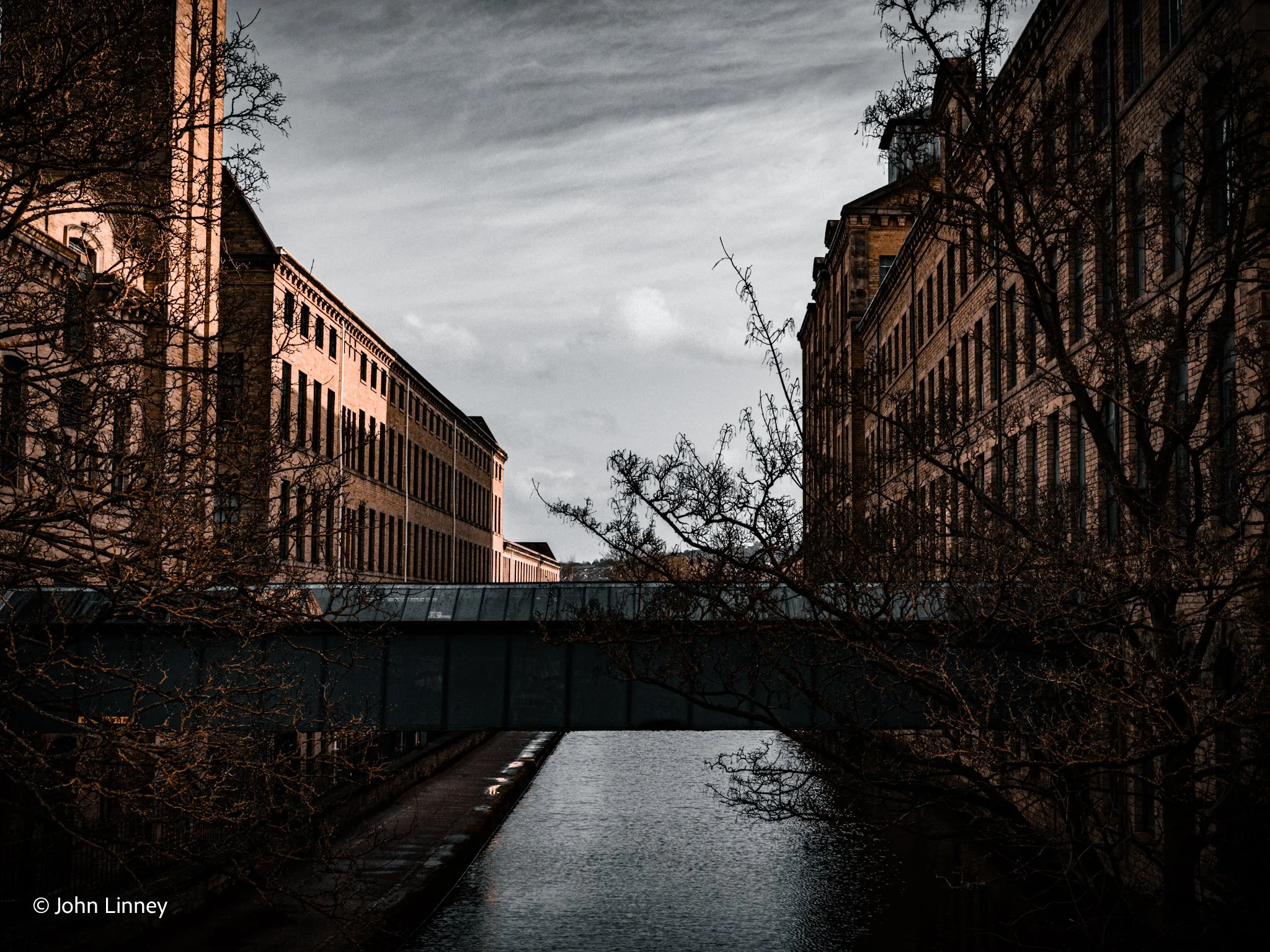 Two historic industrial buildings with a waterway and a footbridge between them, framed by bare trees.