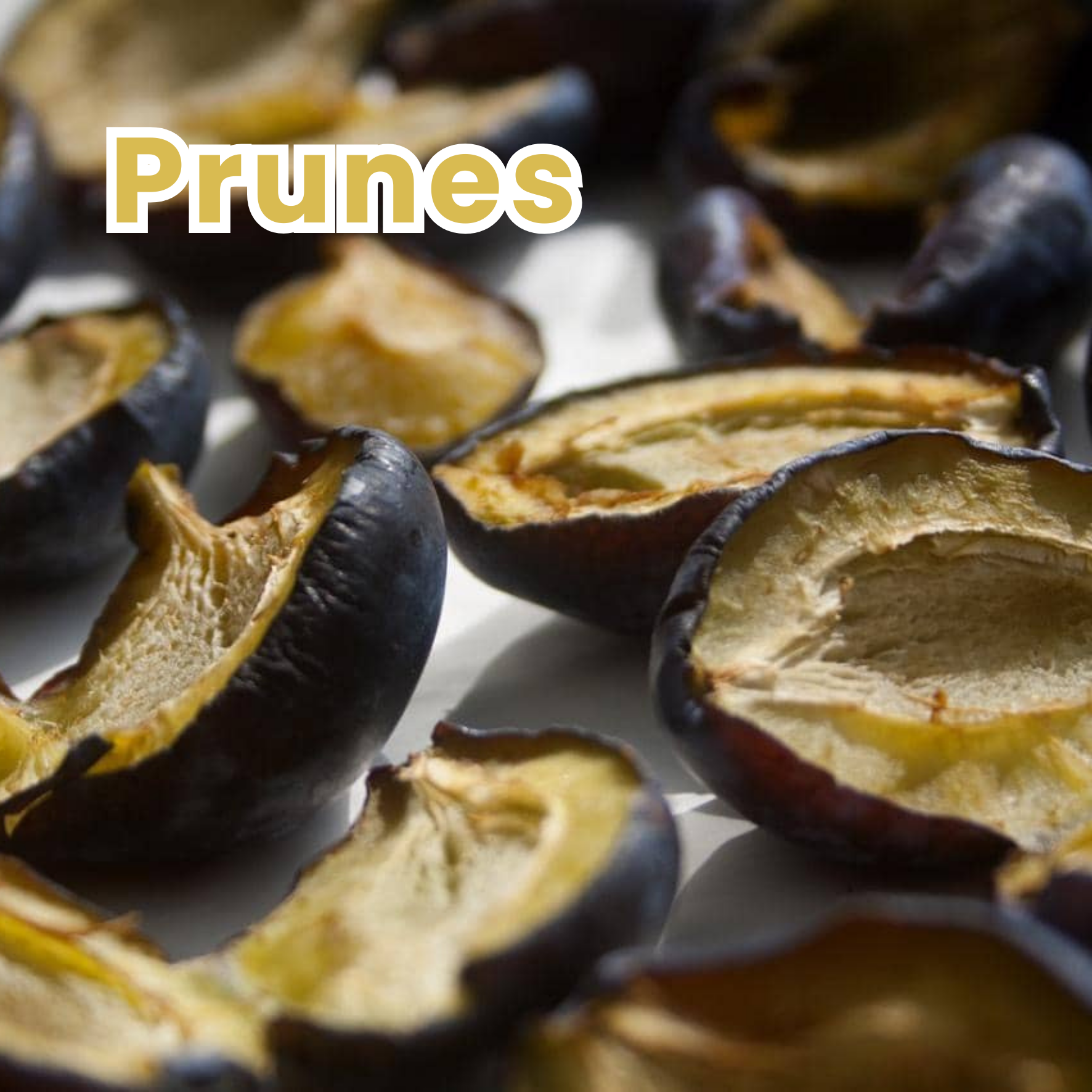 Close-up of roasted prunes on a white surface with the label "Prunes" in bold, tan-colored text.