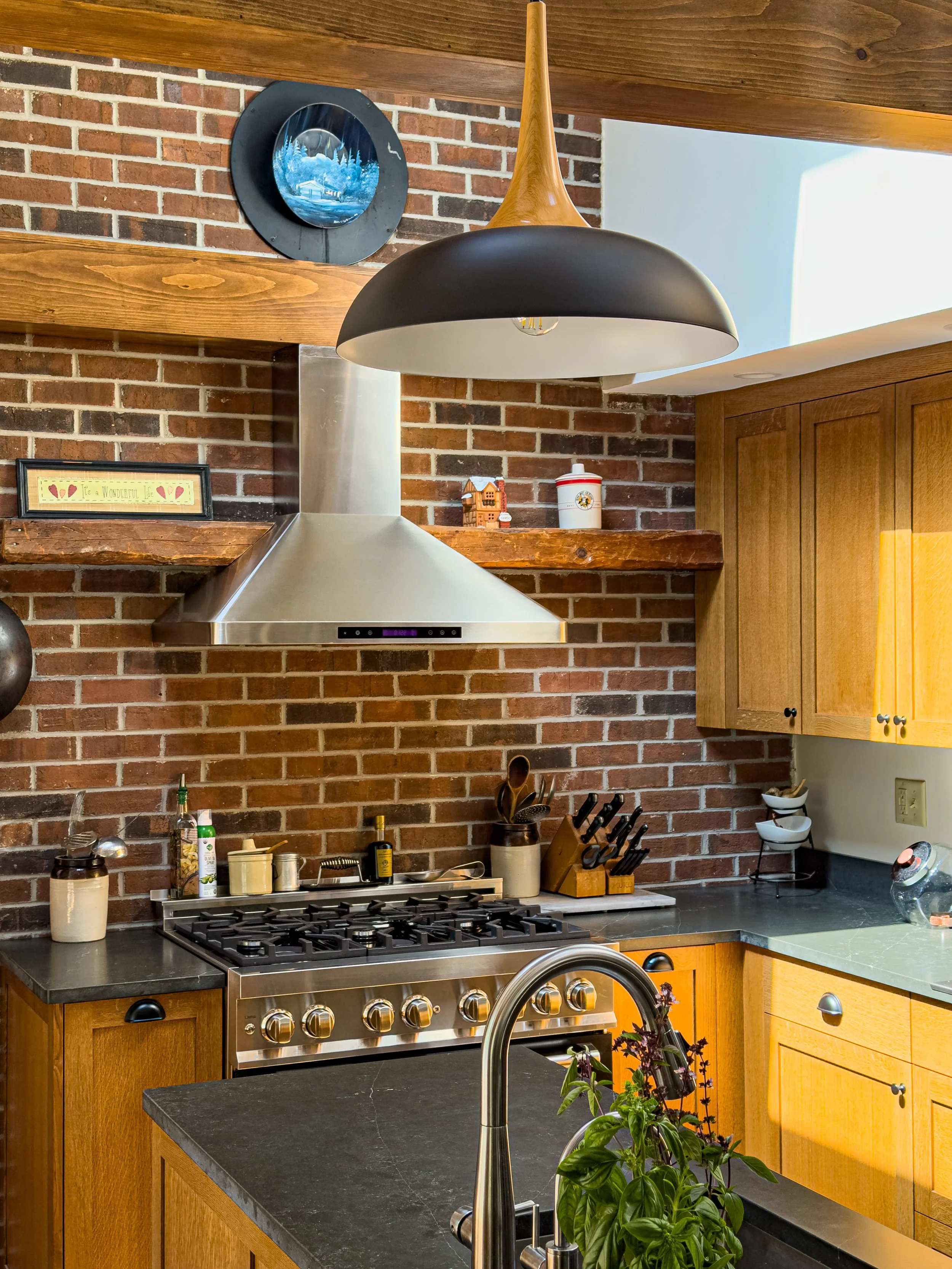 Kitchen with brick wall, black countertop, wooden cabinets, stainless steel stove, range hood, a faucet, potted plant, and decorative items on a wooden shelf.