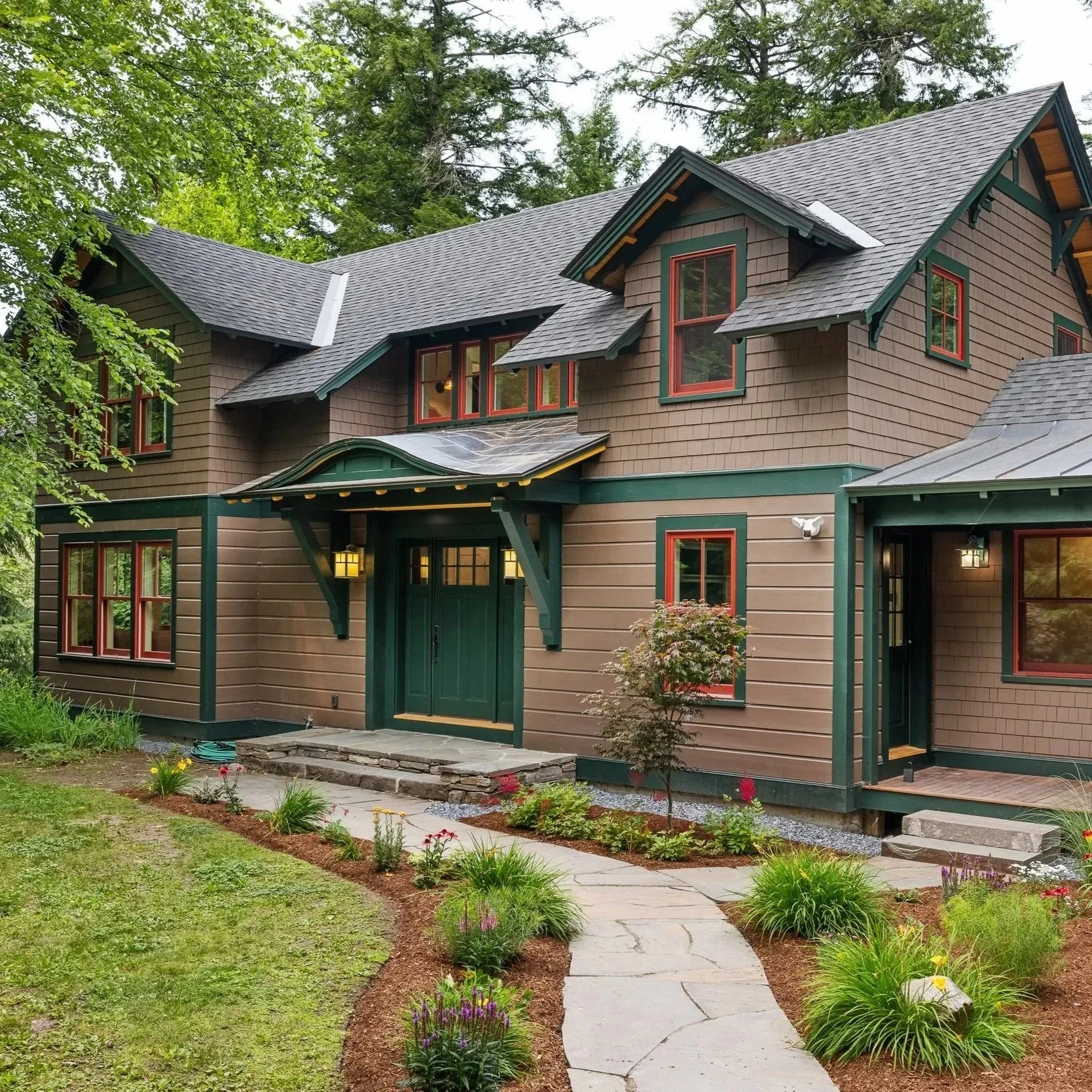 A two-story house with brown shingle siding, green trim, and multiple gabled roofs surrounded by a landscaped yard with flowers, plants, and a curved stone pathway.