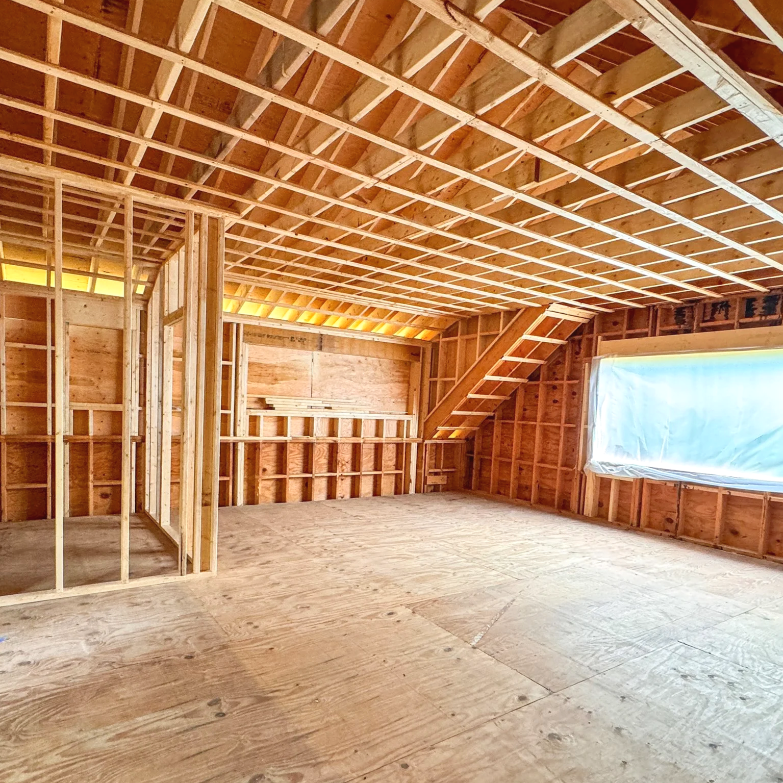 Interior of a house under construction showing wood framing, exposed ceiling, and a window covered with a plastic sheet.