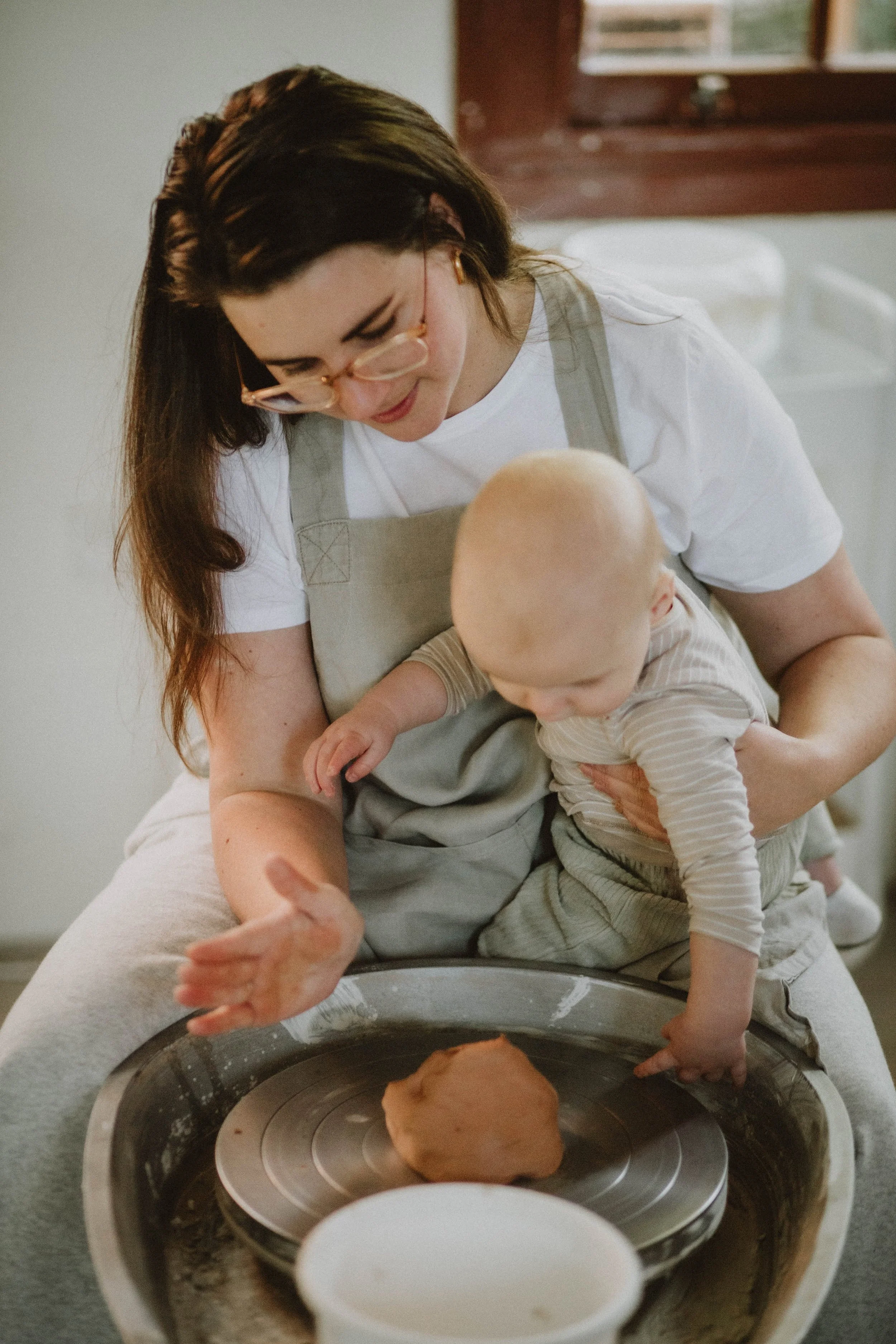 A woman with long brown hair and glasses kneels on a pottery wheel while holding a baby. The woman is smiling as she guides the baby's hand on the clay. The baby is reaching out to touch the clay on the wheel. There is a small bowl of water in front of the wheel, and a window with brown trim in the background.