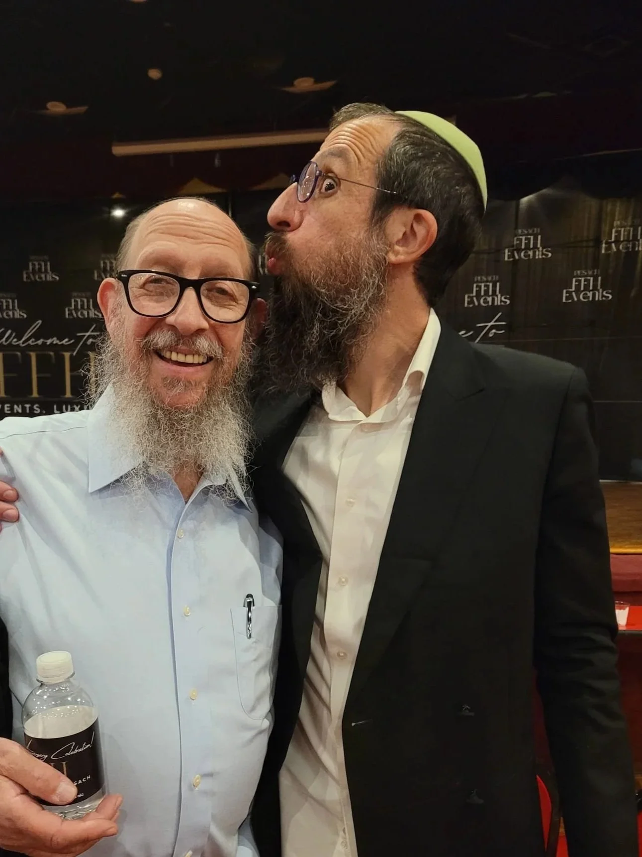 Two men, one with a gray beard and glasses, smiling, holding a water bottle, and the other with a longer beard, glasses, and a green skullcap, kissing on the cheek at an event with a black backdrop.