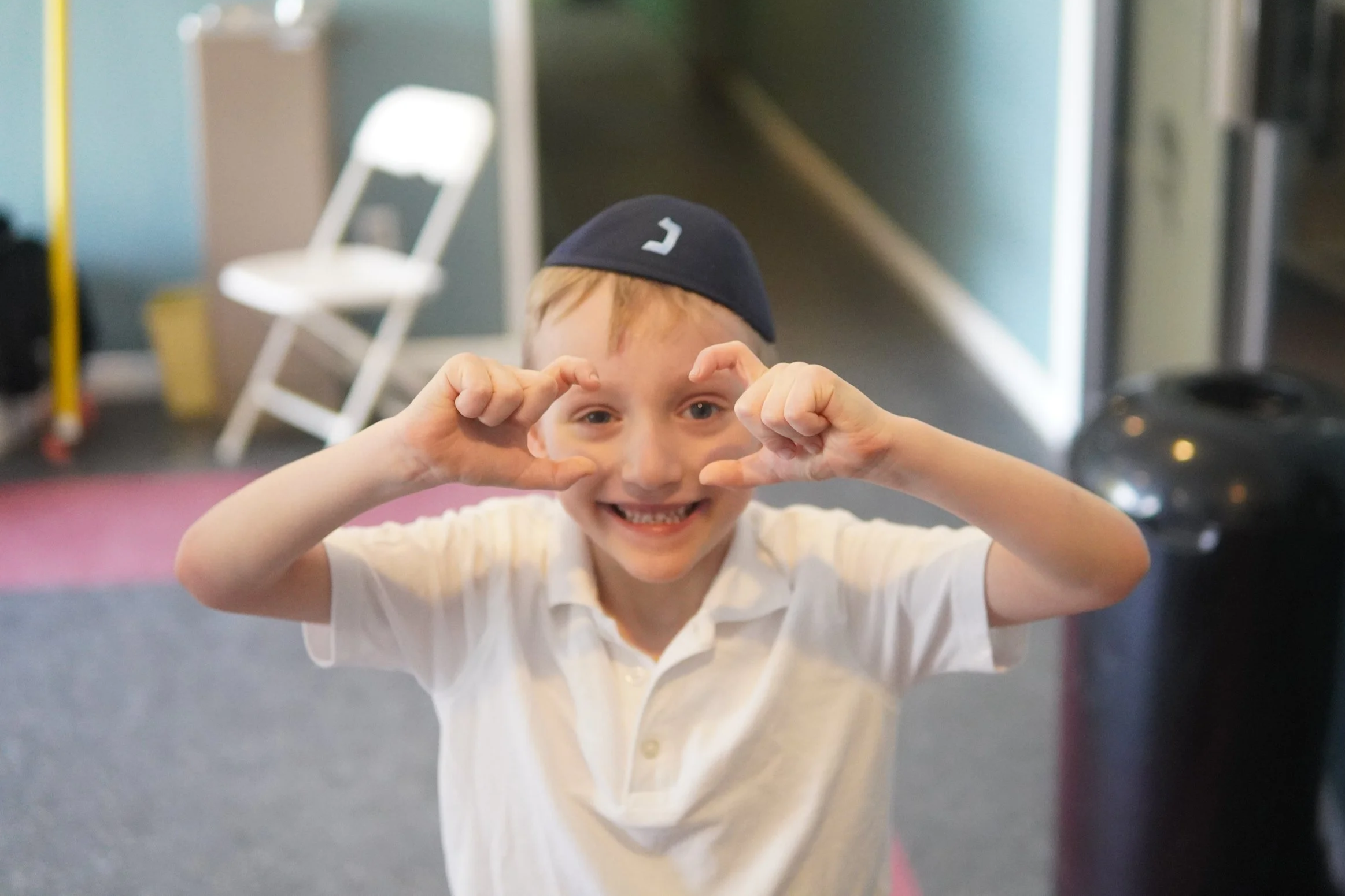 A young boy wearing a black cap and white polo shirt smiling and making a framing gesture with his hands in front of his face.
