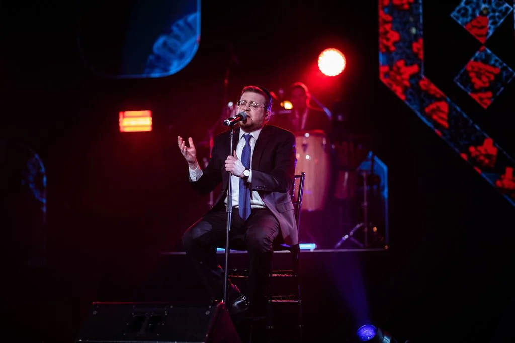 A man in a suit singing into a microphone on stage with colorful lighting and a musician playing percussion in the background.