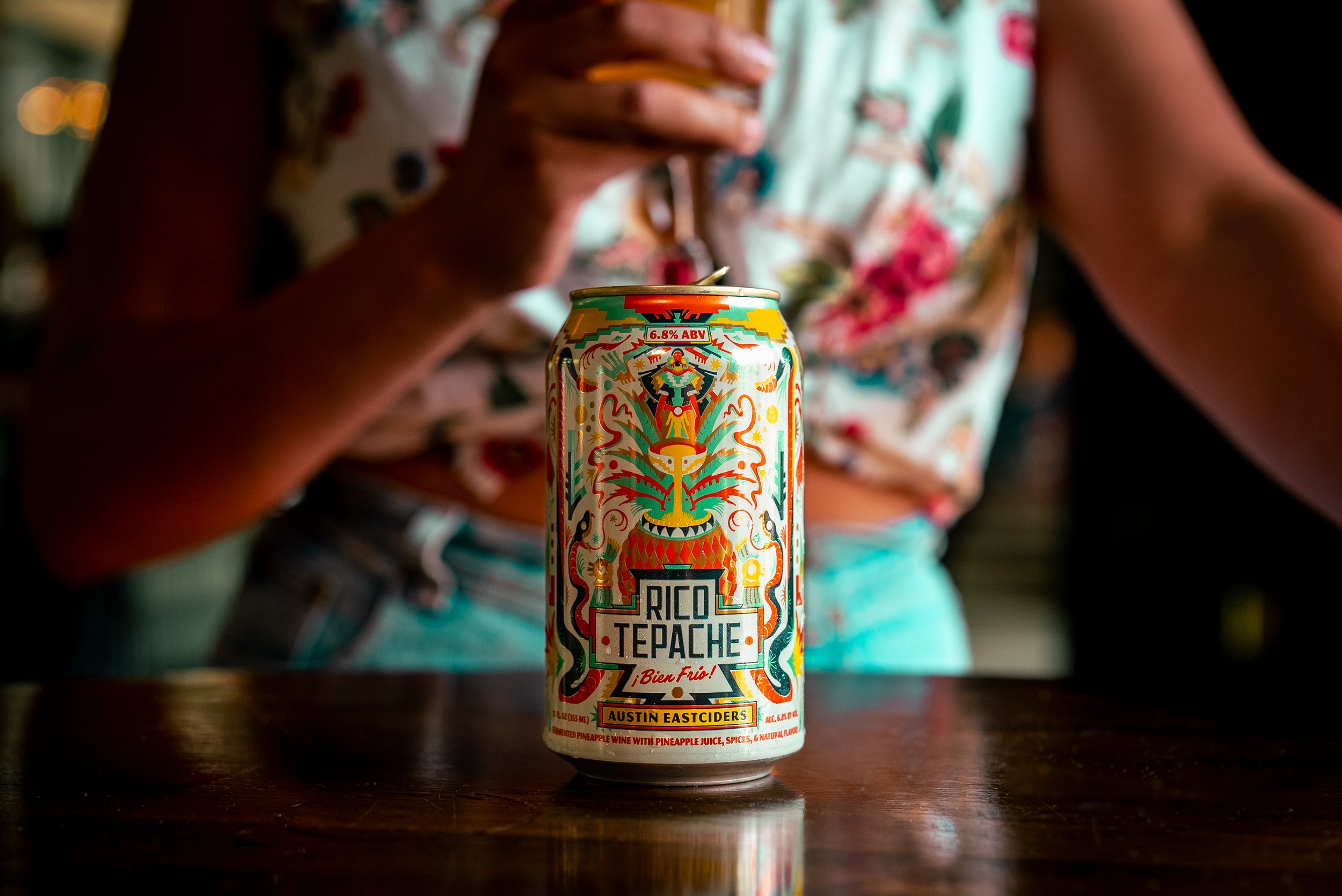 Commercial beverage and food product photography of a Rico Tepache can by Austin Eastciders on a wooden surface, featuring vibrant colors and a lifestyle setting with a softly blurred figure in the background, creating an eye-catching, high-impact im