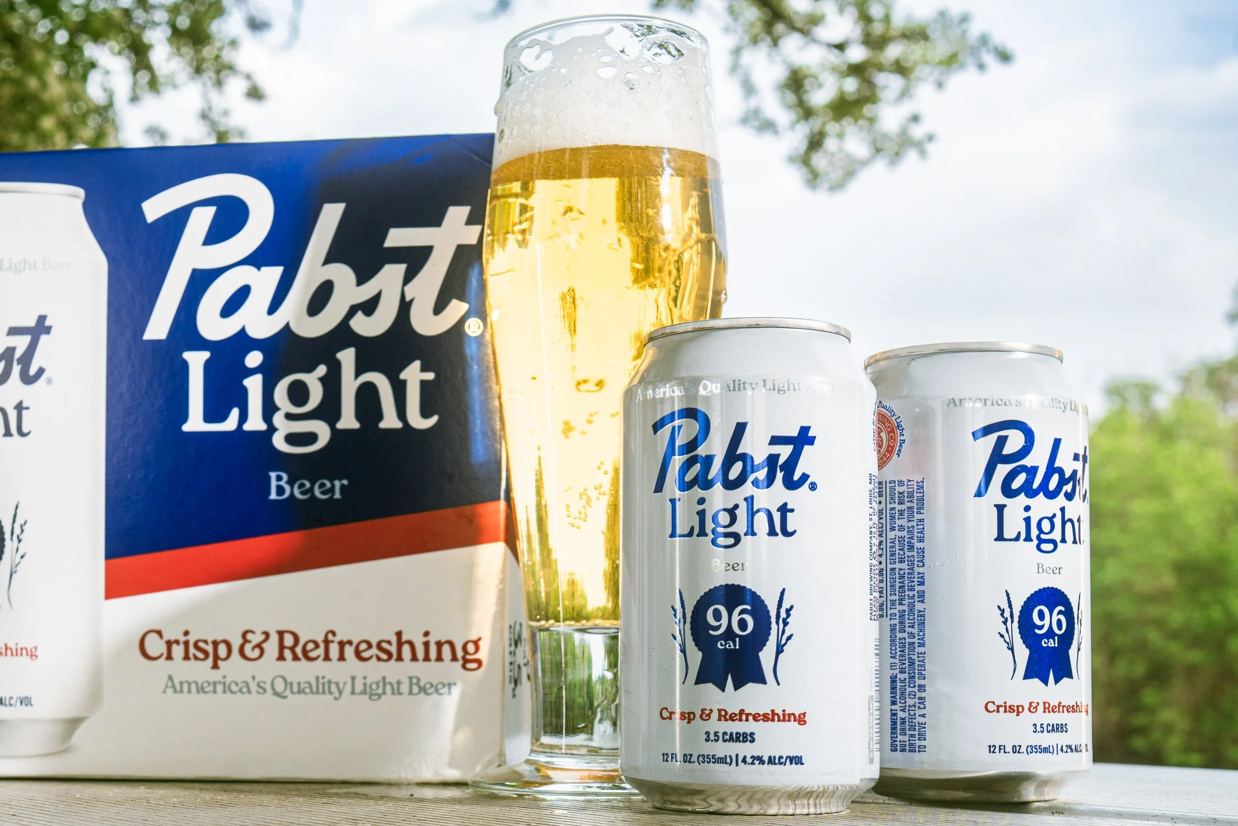 Commercial beverage and food product photography of Pabst Light beer with packaging, cans, and a poured glass on a wooden surface in an outdoor lifestyle setting for social media and advertising content in Austin, Texas.