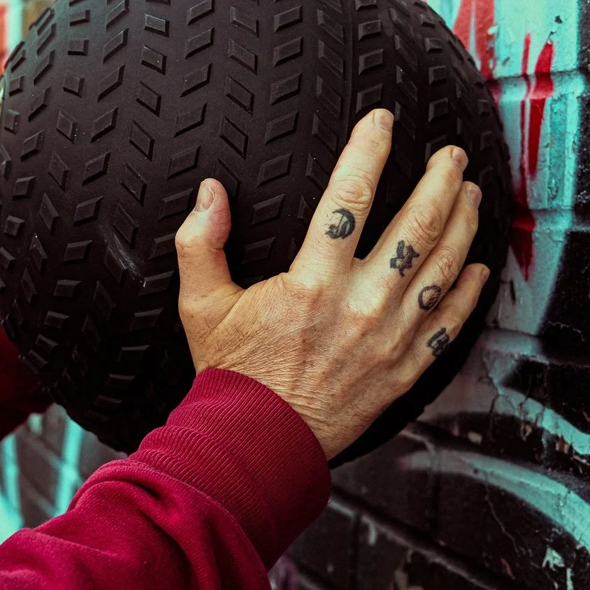 A person's hand with tattoos on fingers, wearing a red sleeve, resting on a black tire against a graffiti-covered wall.