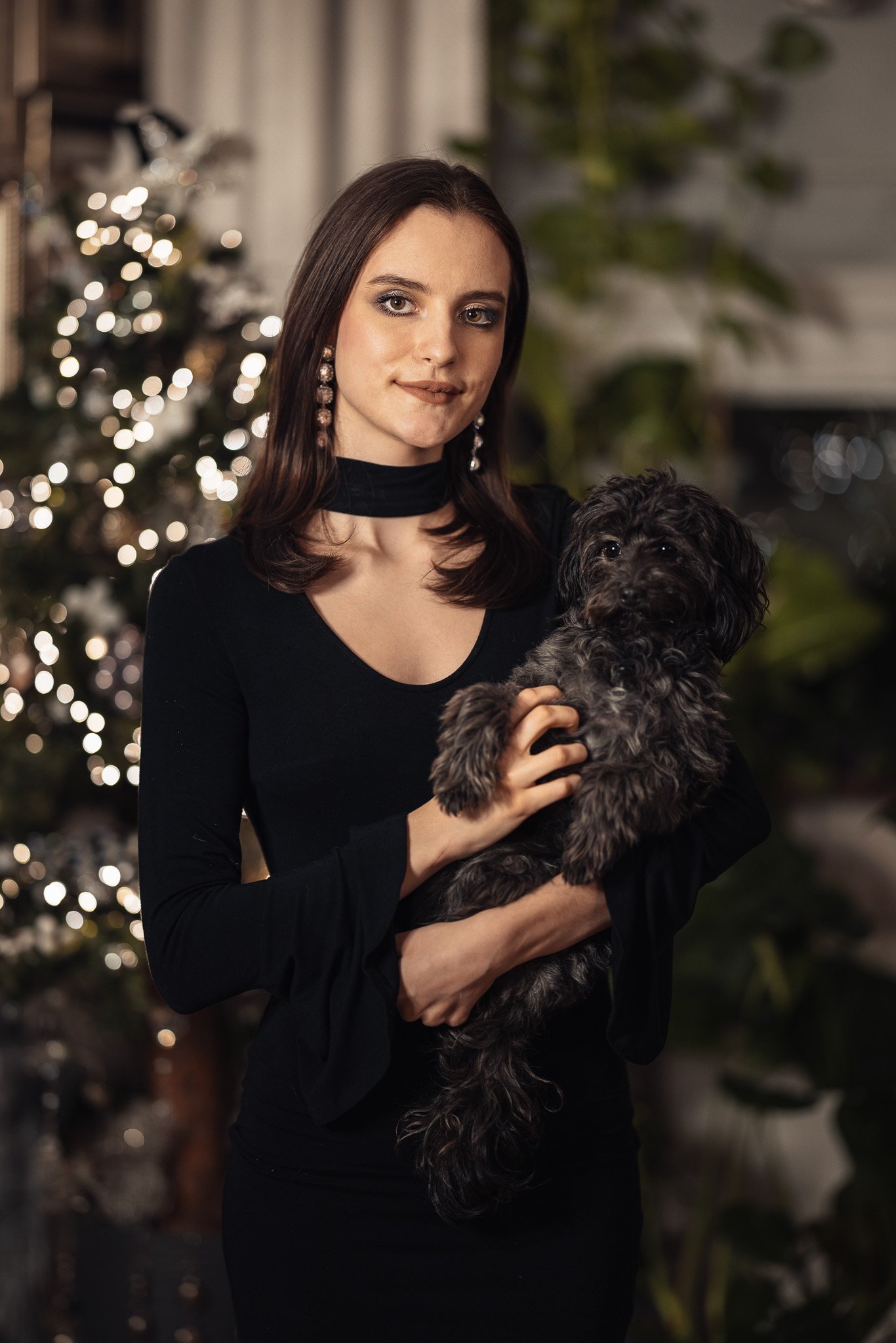 Kvinne i svart kjole holder en liten, grå hund foran et pyntet juletre.