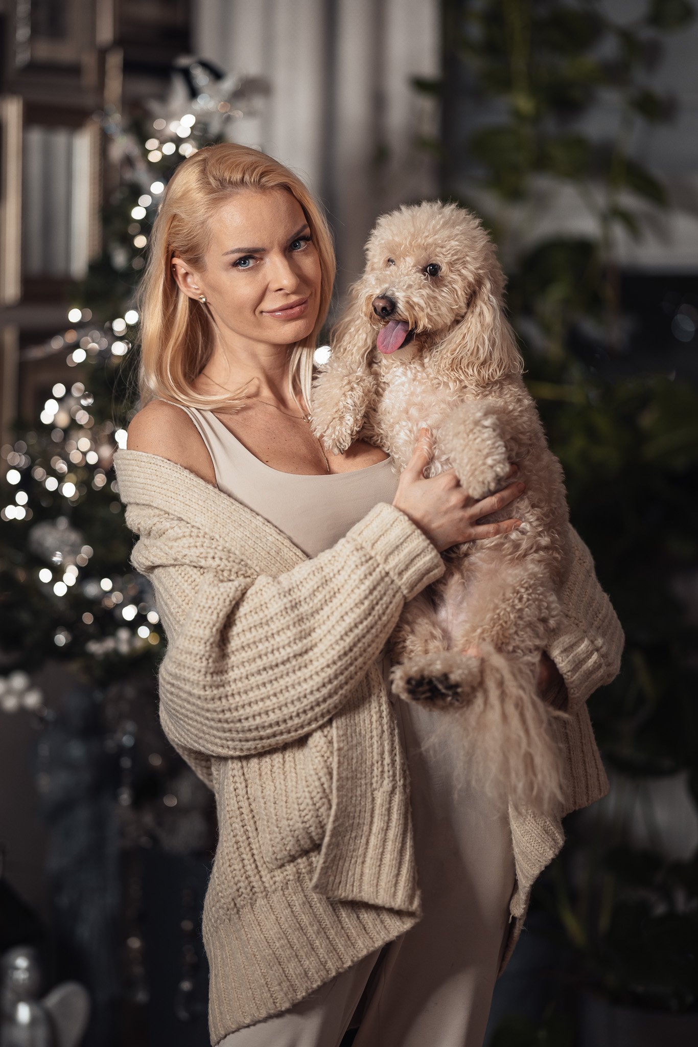 Kvinne med blondt hår i lys cardigan holder en liten, fluffy hund foran et dekorert juletre.