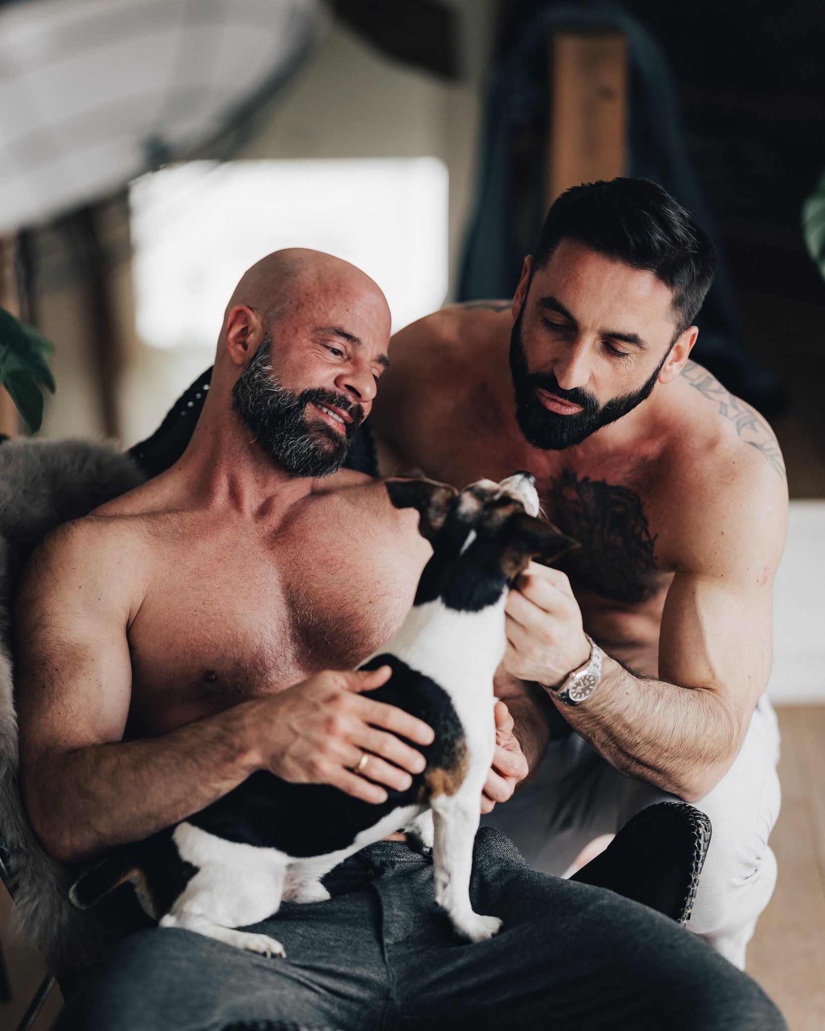 To skjeggete menn uten skjorter sitter sammen og leker med en liten hund i fanget. De ser glade ut og har øyekontakt med hverandre og hunden.
