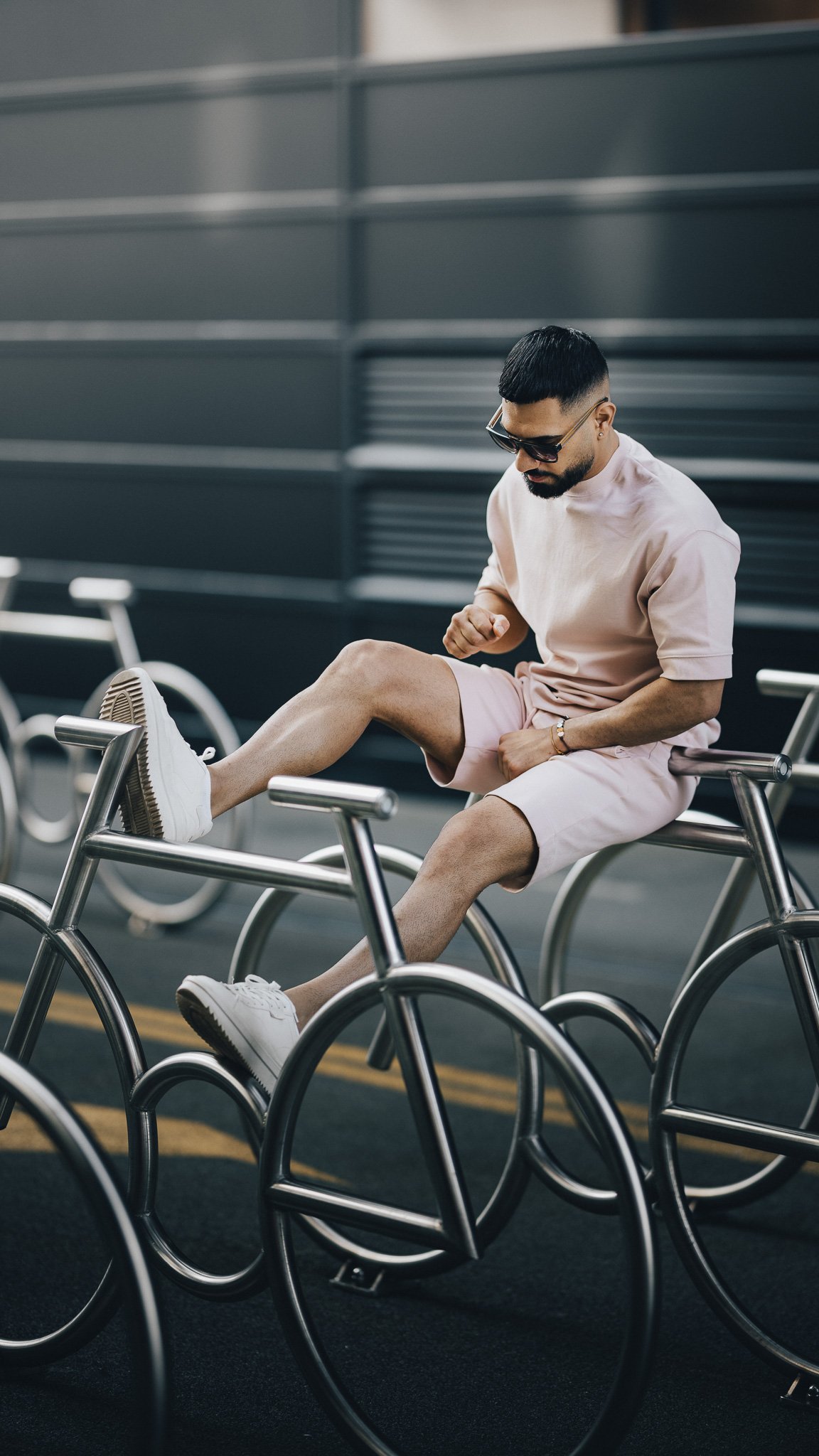 Mann i rosa antrekk sitter på metallsykkelstativ, iført solbriller og hvite joggesko.