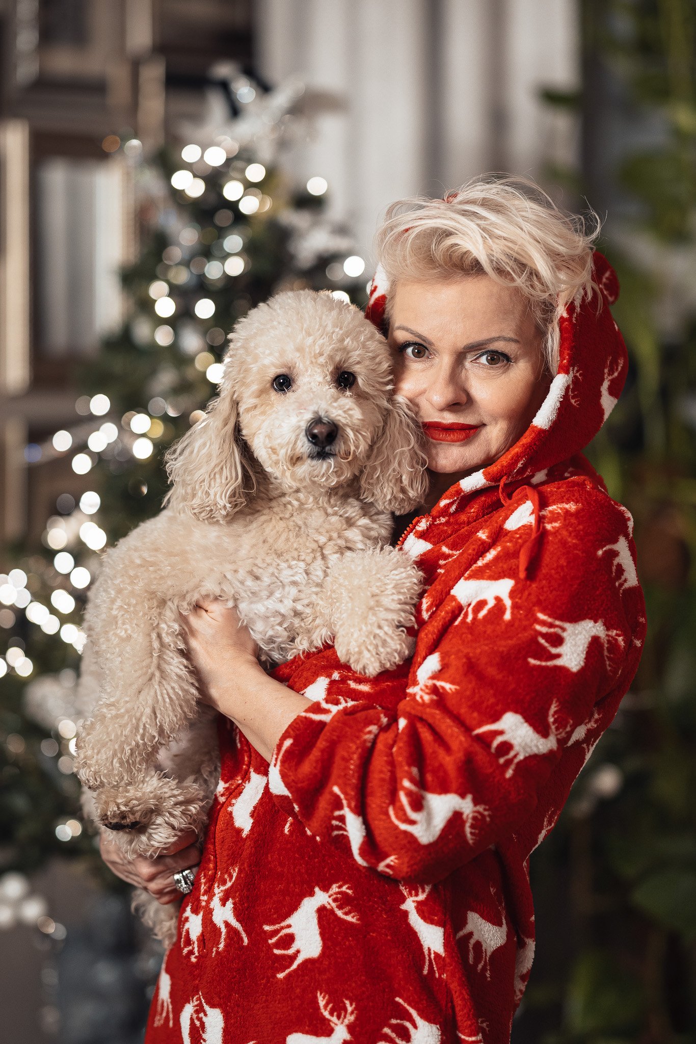 Person i rød hettegenser med reinsdyrmotiv holder en liten hvit hund, juledekorasjoner i bakgrunnen.