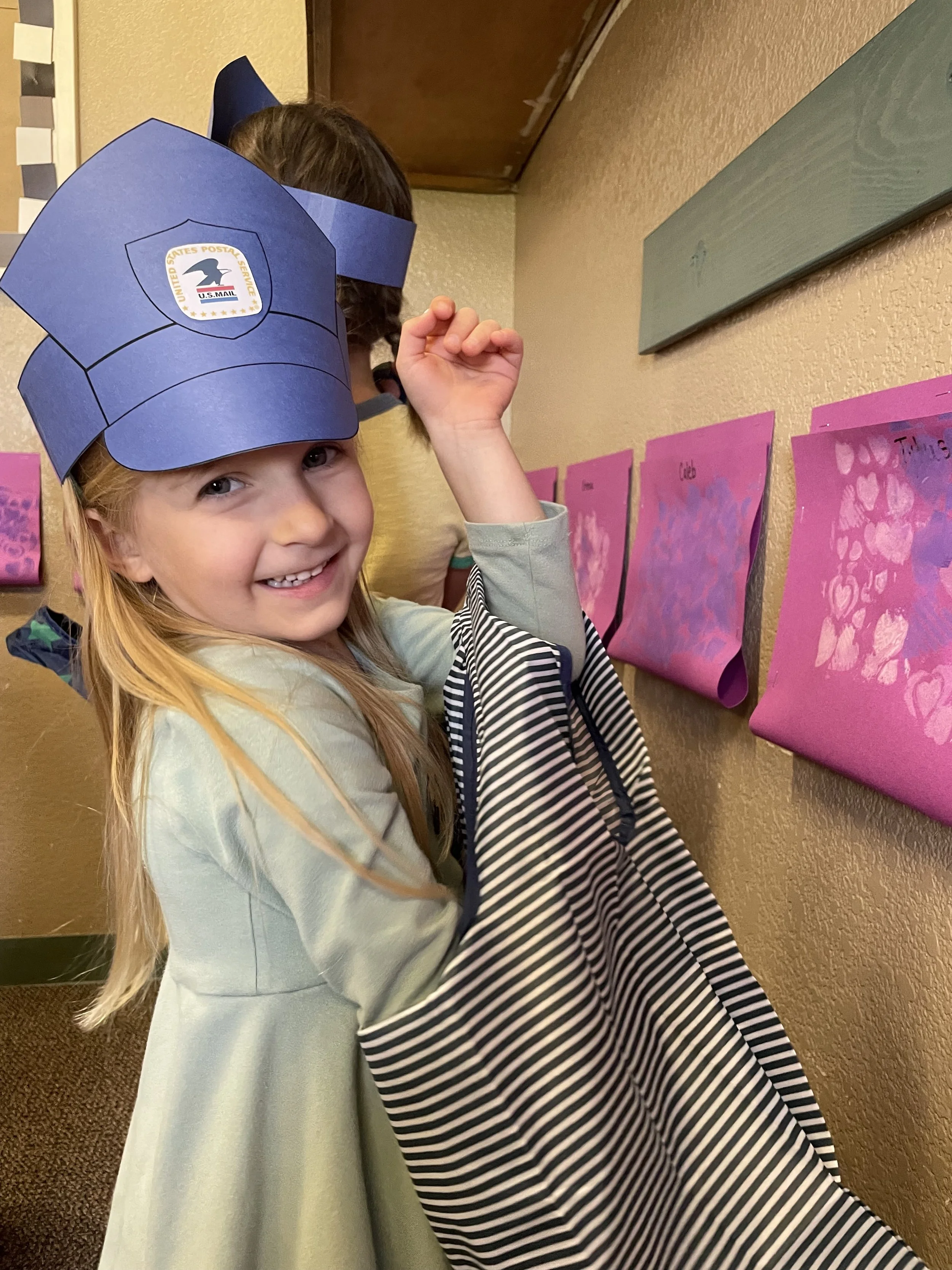 Smiling girl wearing a blue USPS Postal Worker hat and a gray dress, standing next to pink paper rolls with heart-shaped designs on a wall.