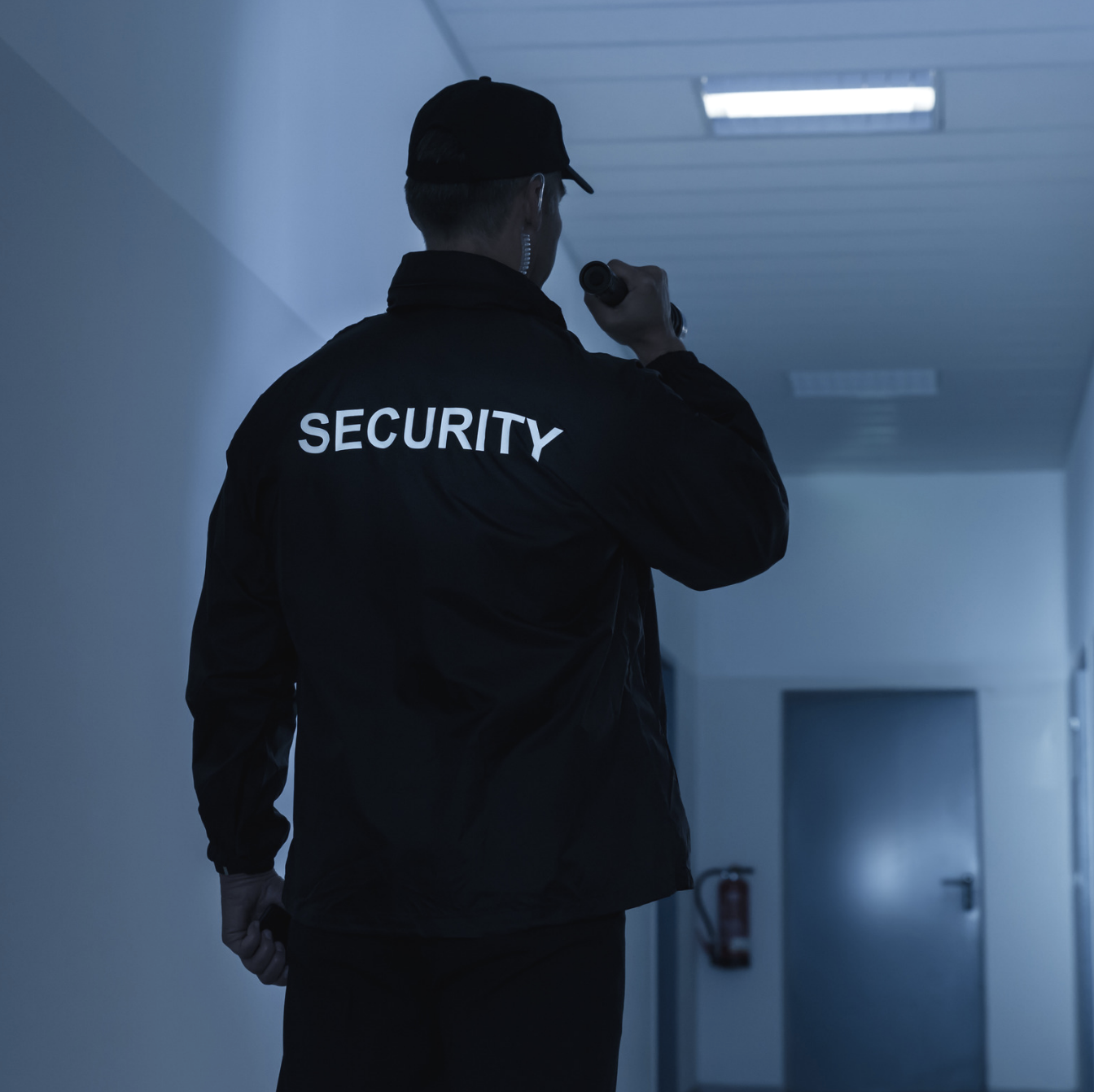 Security guard in black uniform with 'SECURITY' written on back, holding a flashlight, standing in a dimly lit hallway.
