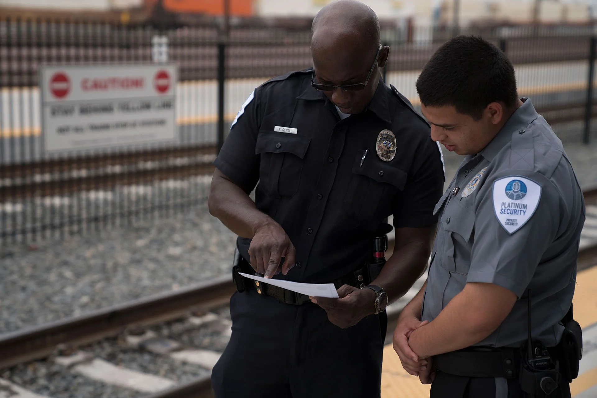 Two police officers, one male and one non-binary, standing on a train platform, discussing something while looking at a paper.