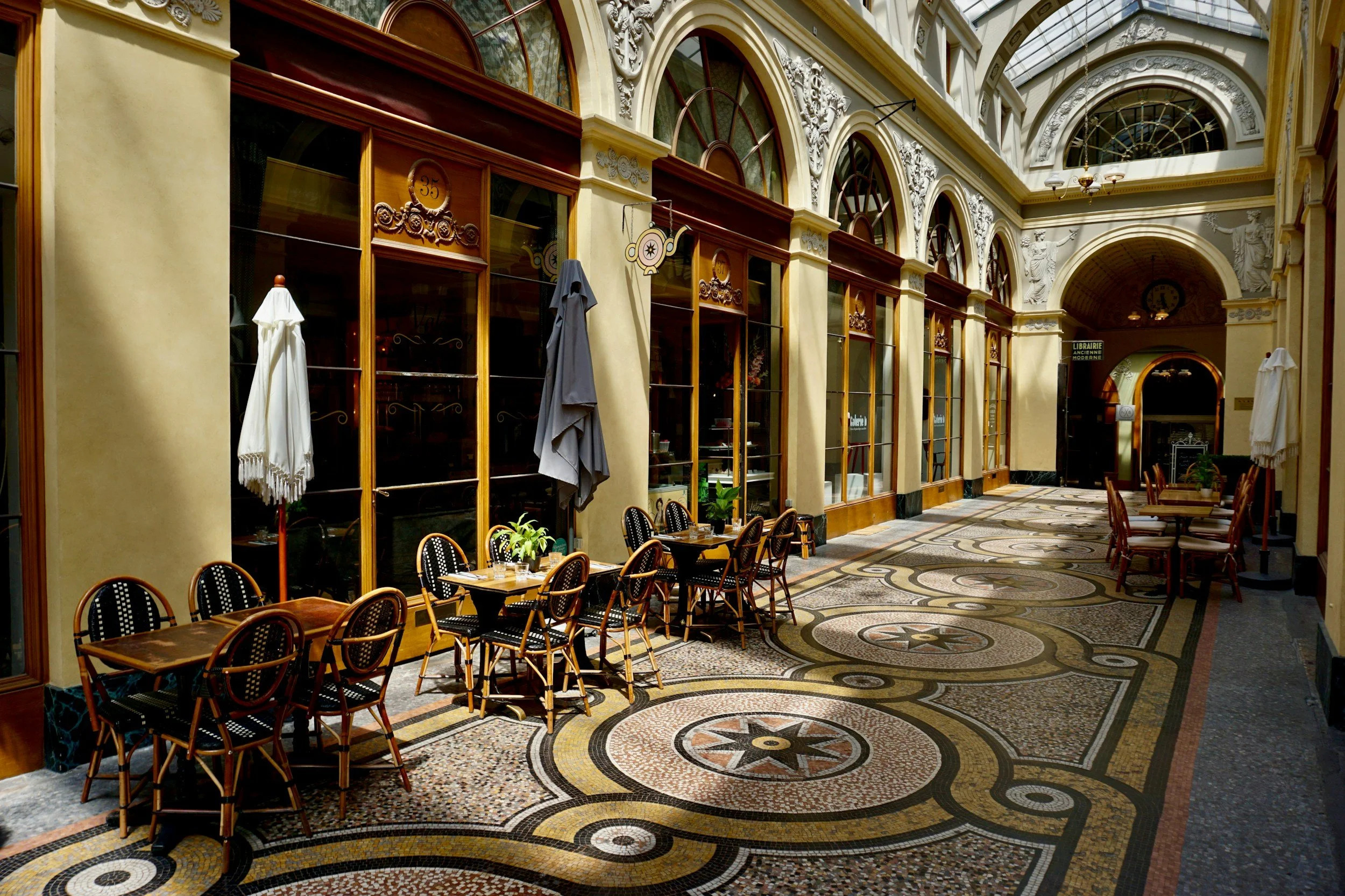 Passage couvert avec verrière, mosaïque au sol et terrasses de café. Photographie d’illustration.
