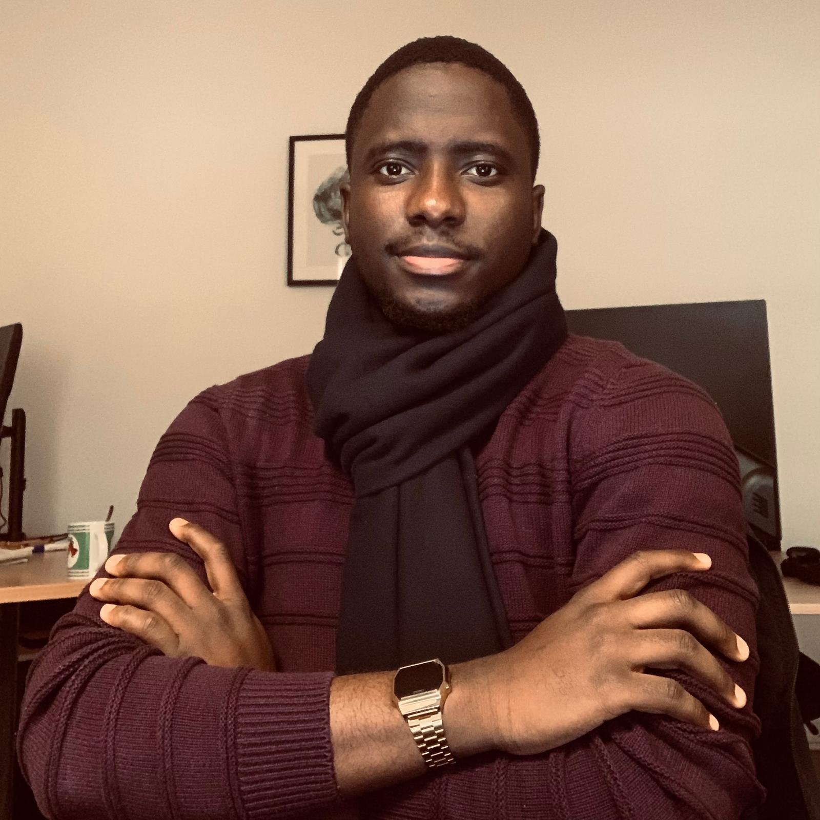 Un homme assis à un bureau avec des bras croisés, portant une veste bordeaux et un foulard noir, dans une pièce avec un ordinateur et une peinture au mur.