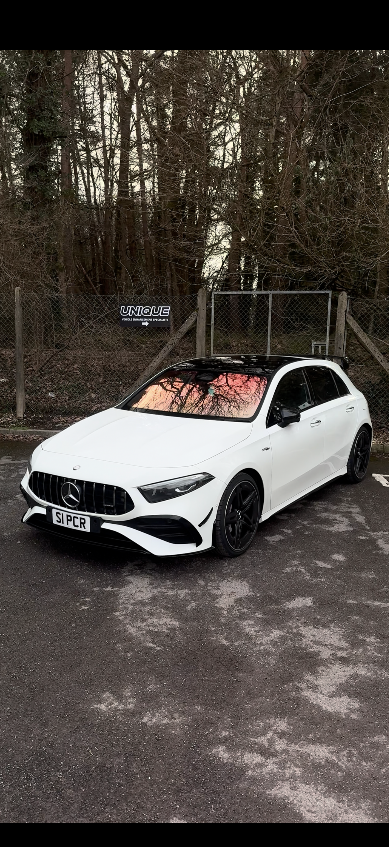 A white Mercedes-Benz car parked in a lot with a black roof and black wheels, in front of a fence with bare trees in the background, during sunset.