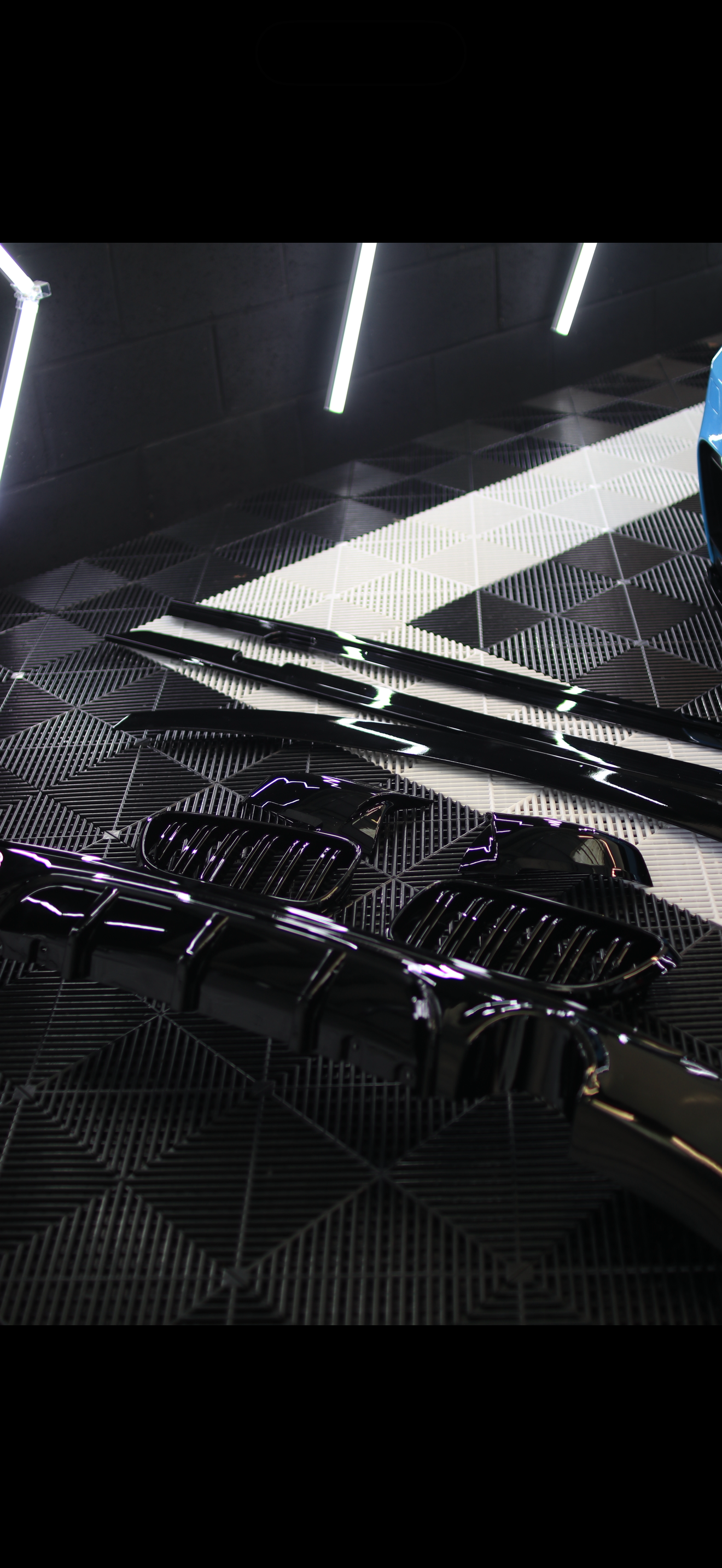 Car parts, possibly a grille or vent, laid out on a patterned surface in a dimly lit environment with black walls and linear white lights overhead.