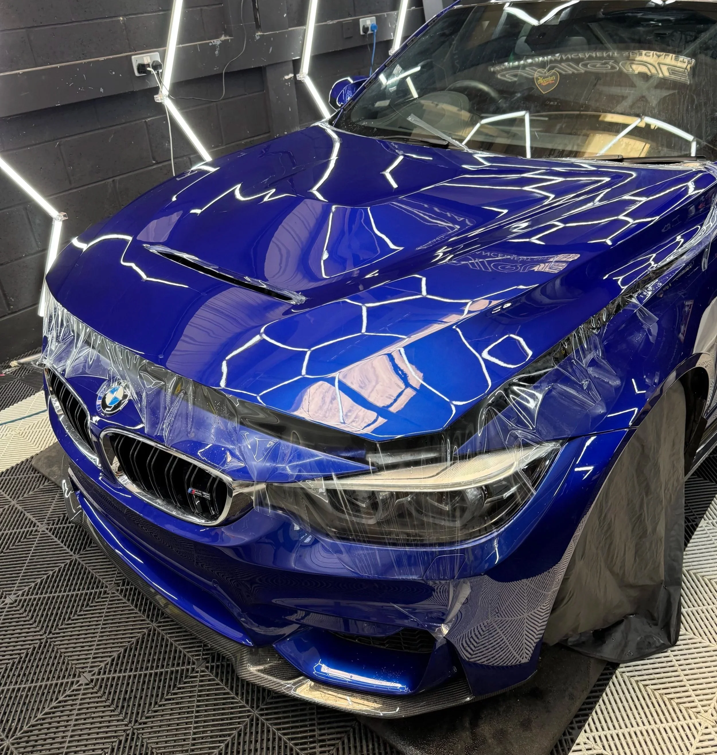 Blue BMW sports car with a sleek, modern design in a garage or workshop, surrounded by reflections from bright, white fluorescent lights on its glossy surface.