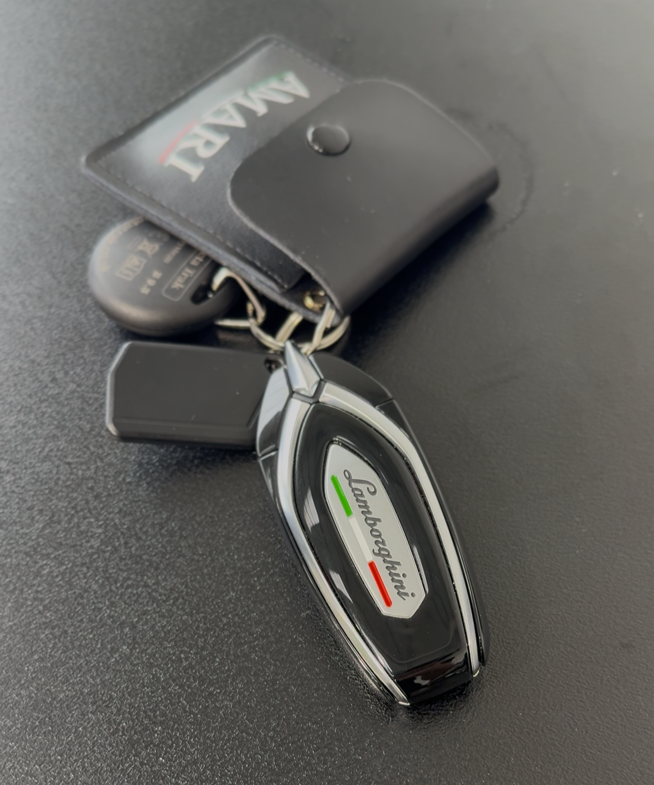 A car key fob with the Lamborghini logo and a black remote, placed on a flat surface, with a car alarm remote nearby.