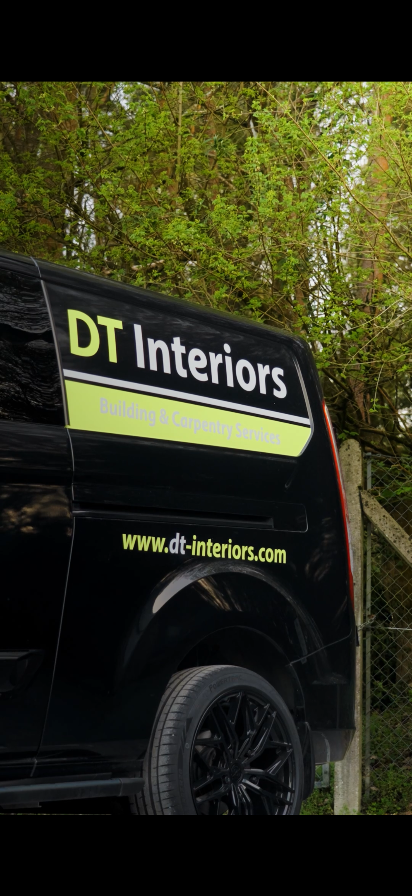 Black van with signage for DT Interiors, a building and carpentry services company, parked outdoors near trees and a fence.