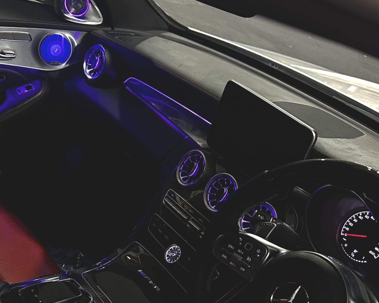 Interior of a luxury car with illuminated purple accents, circular air vents, a digital display, and a steering wheel with controls.