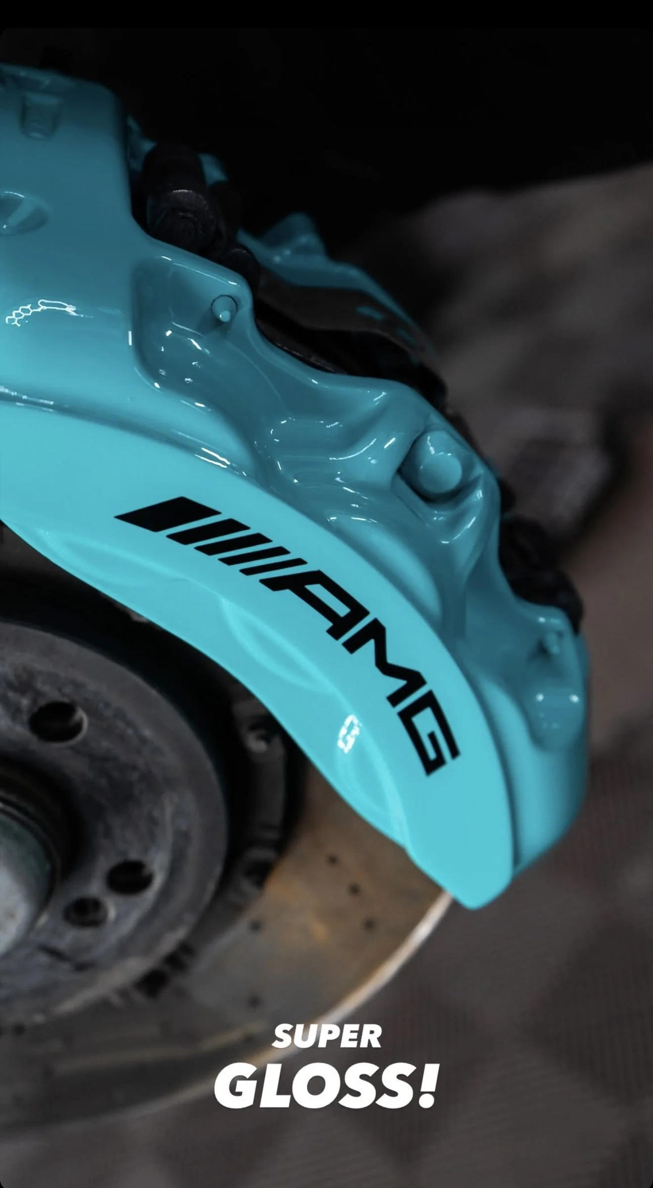 Close-up of a blue motorcycle brake caliper with the logo "AMG" visible, and a blurred brake disc below. The text "SUPER GLOSS!" is overlaid at the bottom of the image.