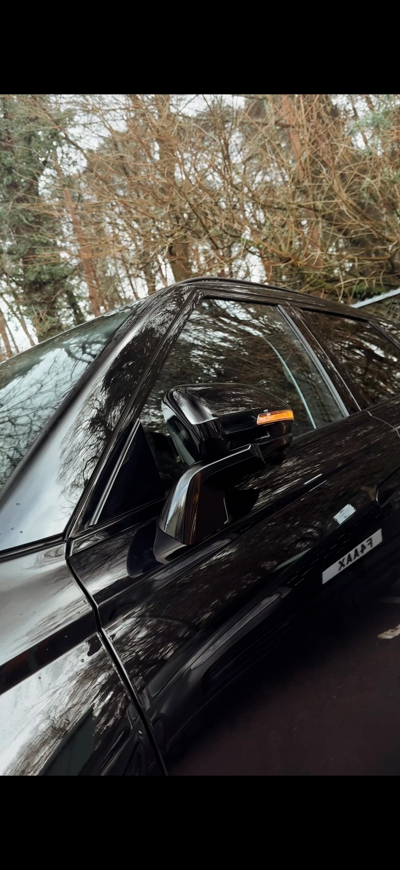 Close-up of a black pickup truck parked outdoors, with trees and branches in the background, reflecting on the shiny surface of the vehicle.