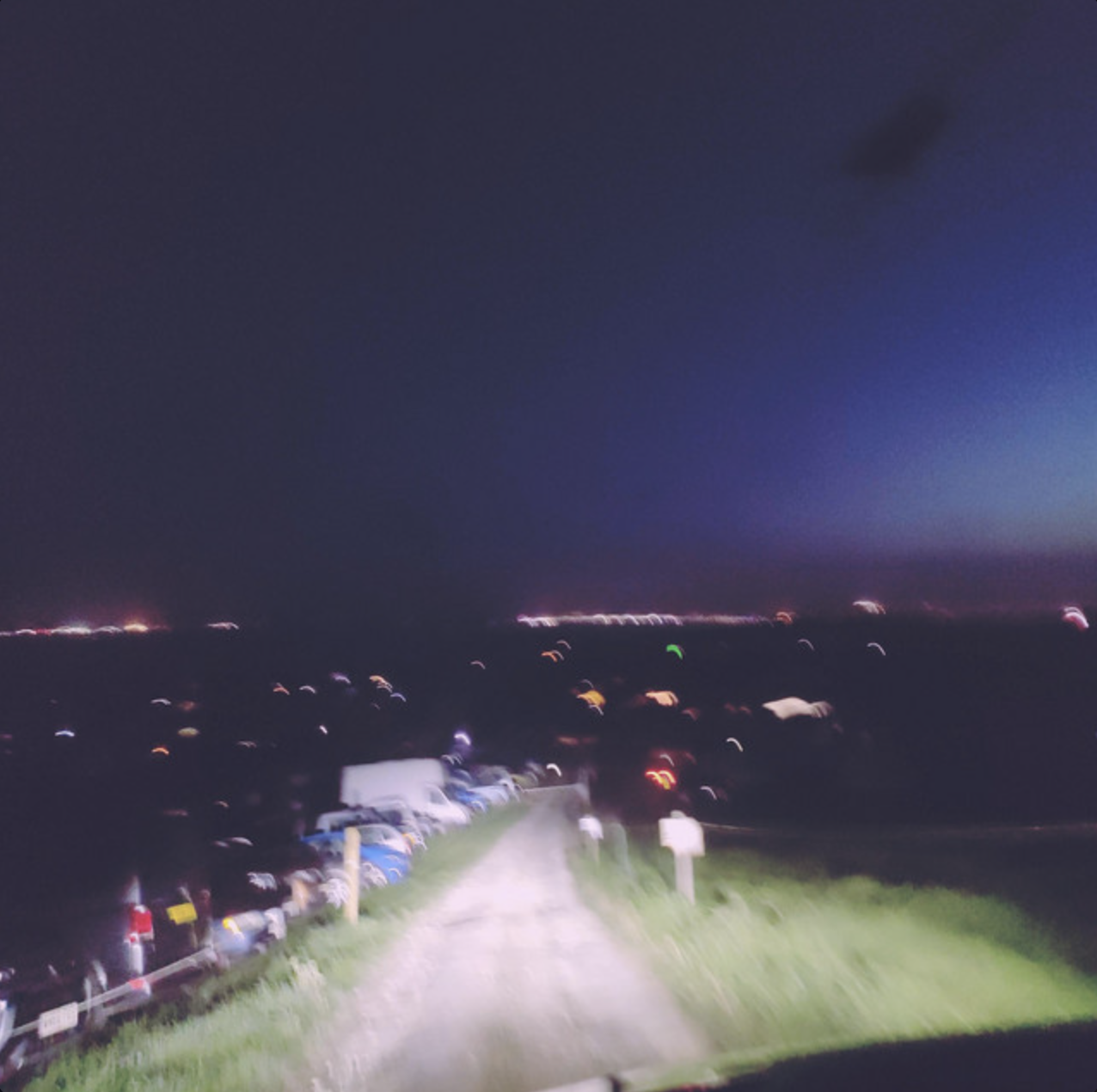 A blurry photo taken at night showing a gravel path with parked cars on the left side, grassy area on the right, and numerous distant city lights under a dark sky.