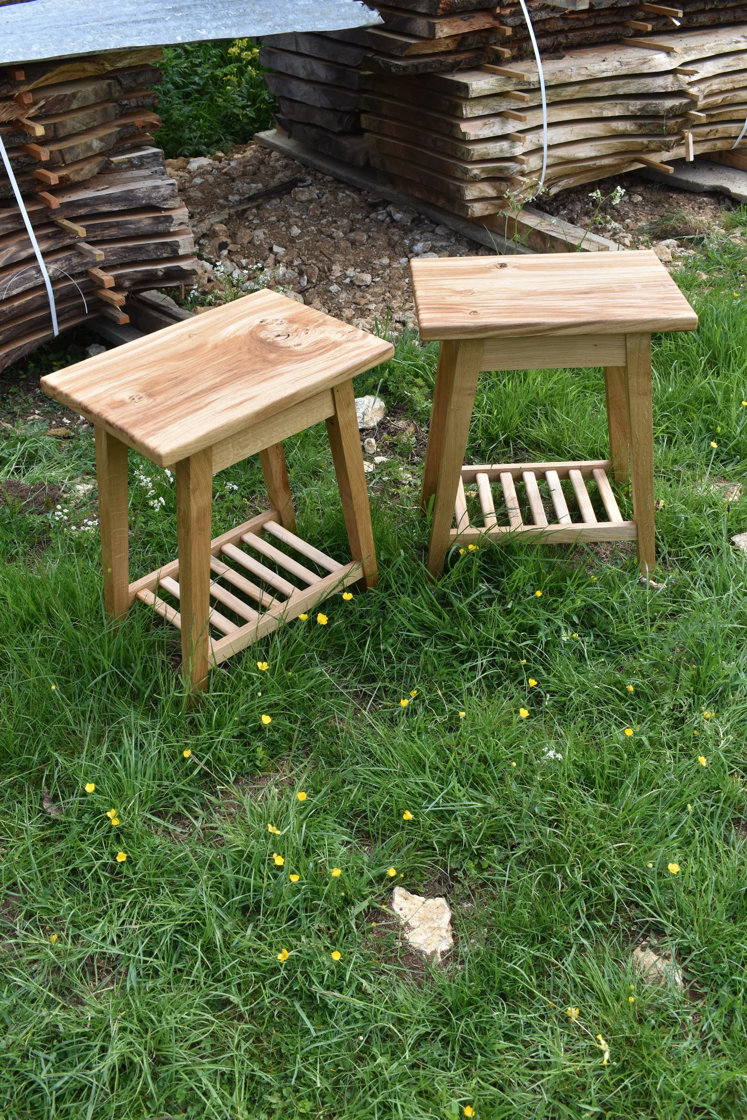 Two wooden stools with slatted lower shelves on green grass with small yellow flowers, near a woodpile.