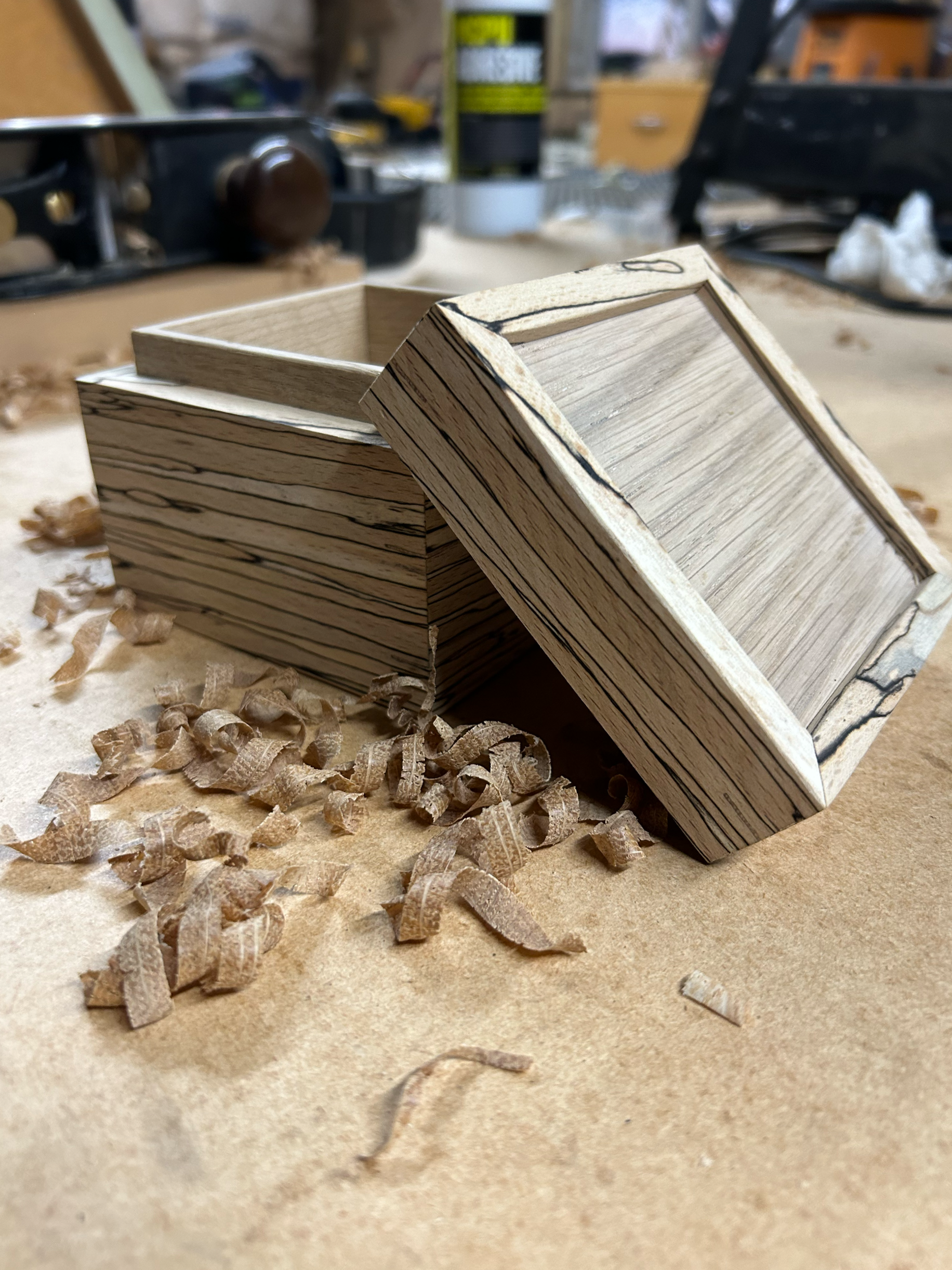 Wooden box with a removable lid, placed on a workbench with wood shavings around it, in a woodworking shop.