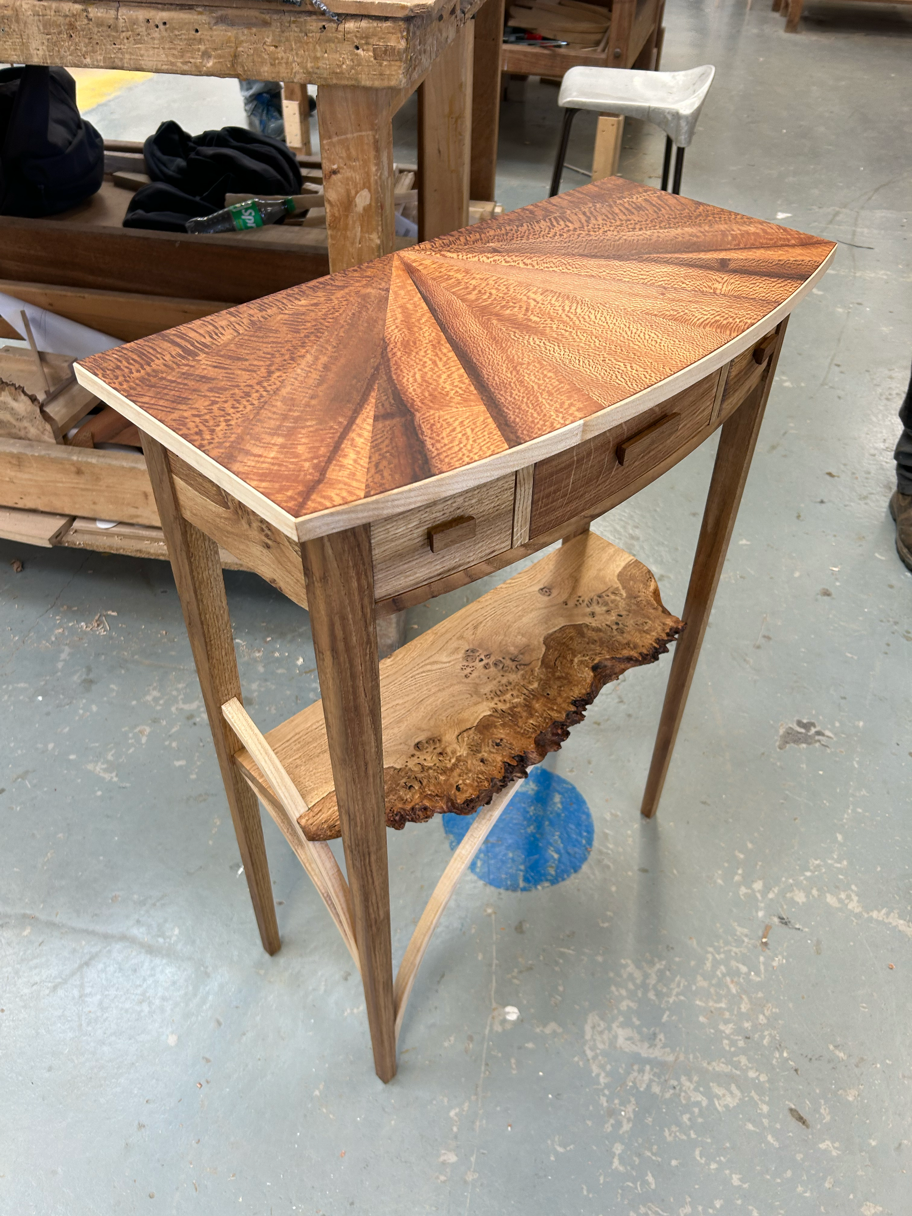 A wooden table with a curved top and an attached drawer, featuring a live edge wood slab on the lower shelf. The table's top has a wood grain pattern, and the space is a woodworking workshop with tools and furniture in the background.