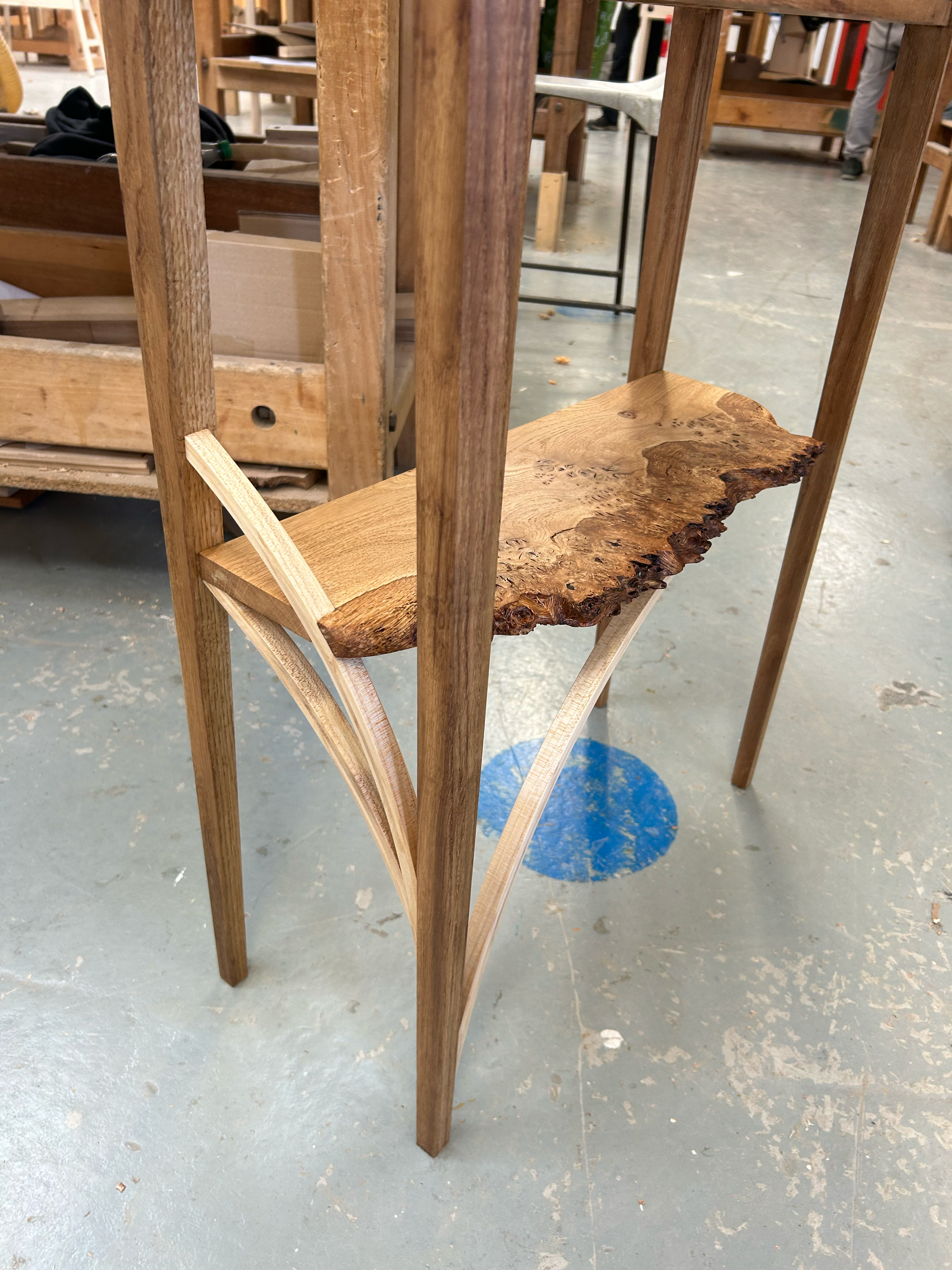 A wooden chair with a natural, uneven wood seat and curved wooden supports, situated on a concrete floor in a woodworking shop.