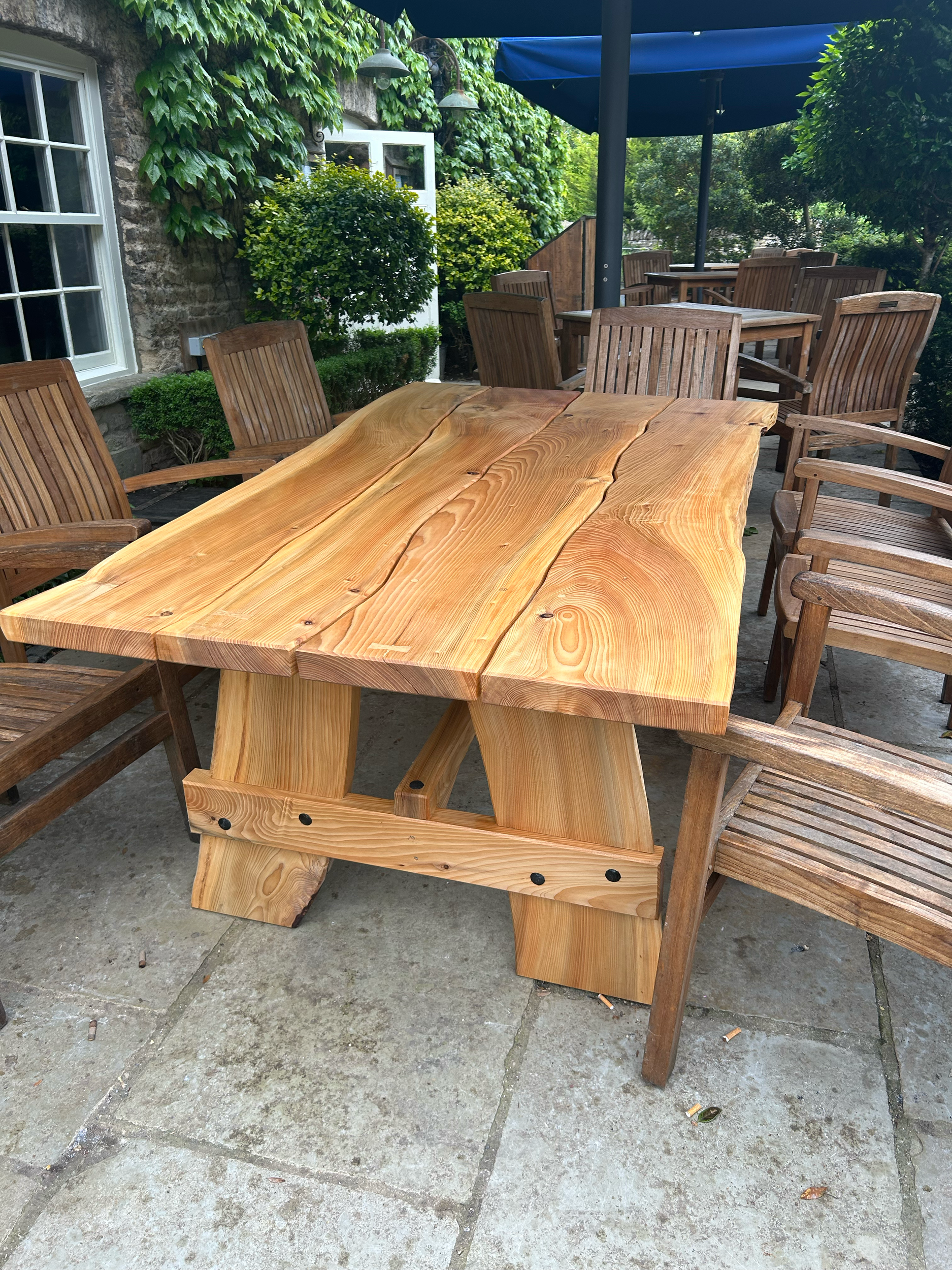 Wooden outdoor table surrounded by wooden chairs with a stone and brick house wall in the background. Green bushes and trees are also visible.