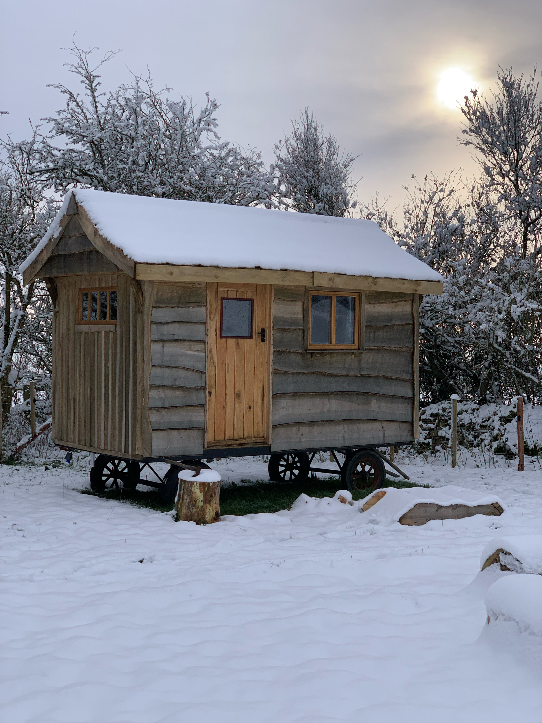 A small wooden house on wheels with a snow-covered roof, windows, and a door, set in a snowy landscape with trees and a cloudy sky, and the sun partially visible behind the clouds.