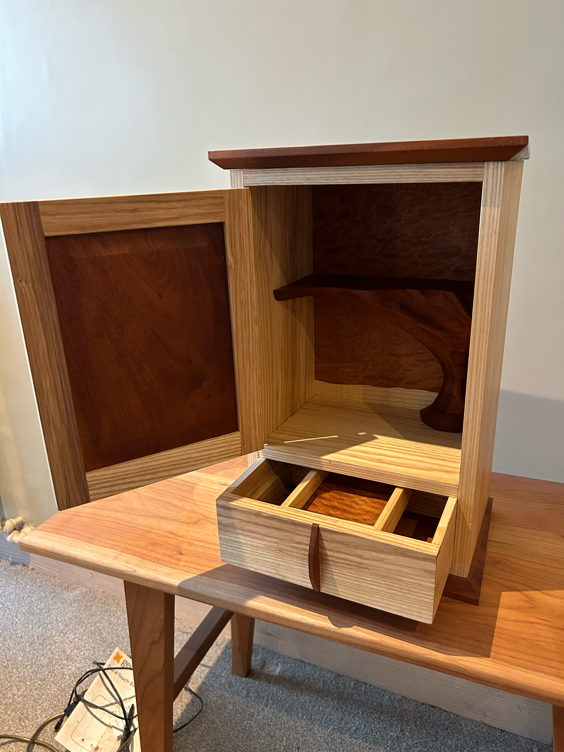 A wooden jewelry cabinet with an open door, inside to the right are shelves, below are small compartments, on the table surface outside is a power strip with cords.